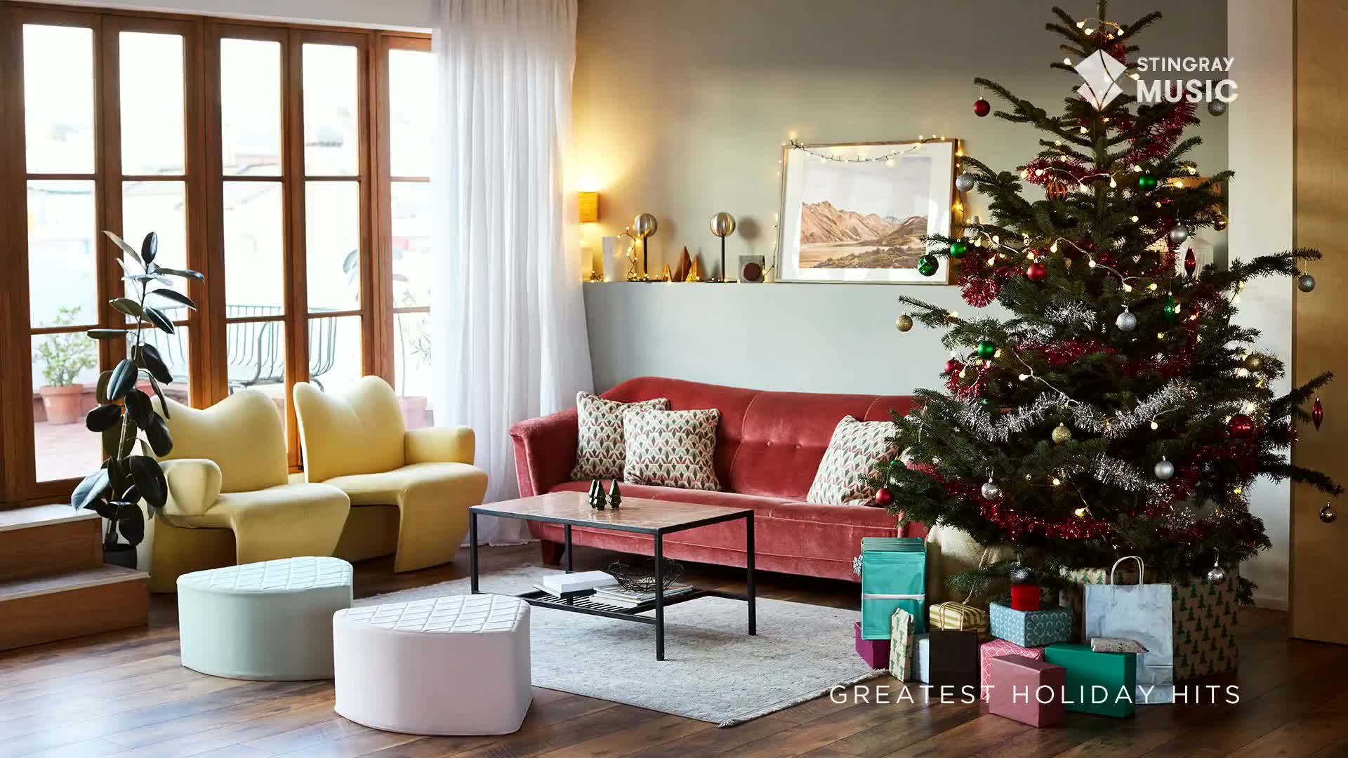 A decorated Christmas tree, with lights and ornaments, stands in the corner of a living room. The scene feels cozy, with a red couch and presents stacked around the tree, fitting for Stingray Holiday Hits from Canada.
