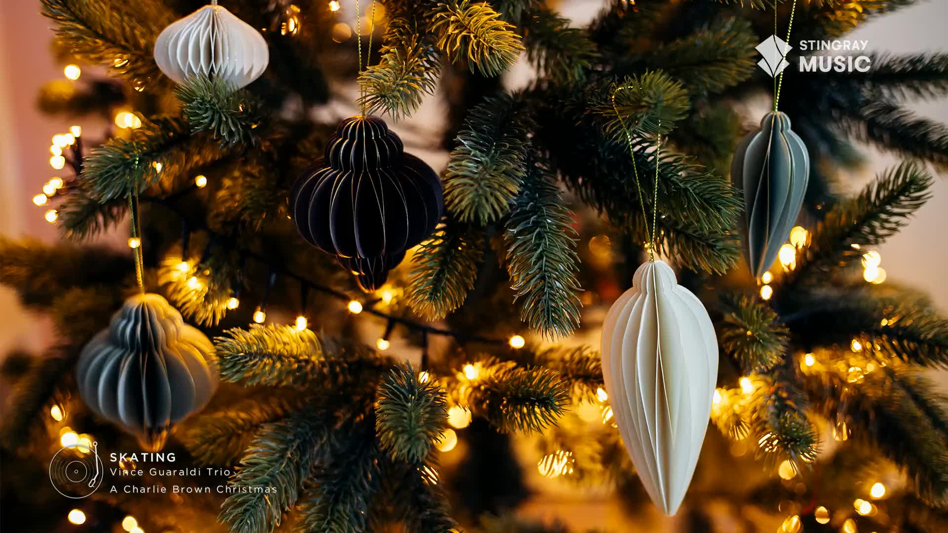 Paper ornaments in shades of white, gray, and black hang from a Christmas tree, illuminated by tiny fairy lights. The Stingray Music logo and the words "Skating" and "A Charlie Brown Christmas" are visible at the bottom.
