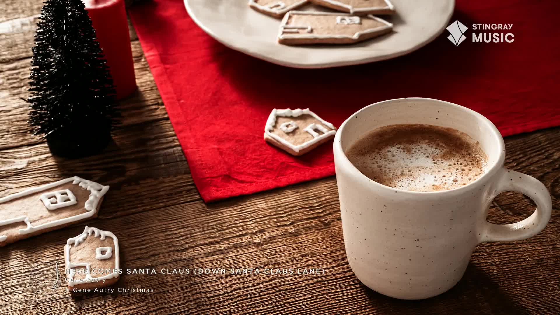 A steaming mug sits beside gingerbread house cookies, the scene set on a wooden surface with a red cloth. The Stingray Holiday Hits logo is visible, and "Here Comes Santa Claus" plays softly in the background.
