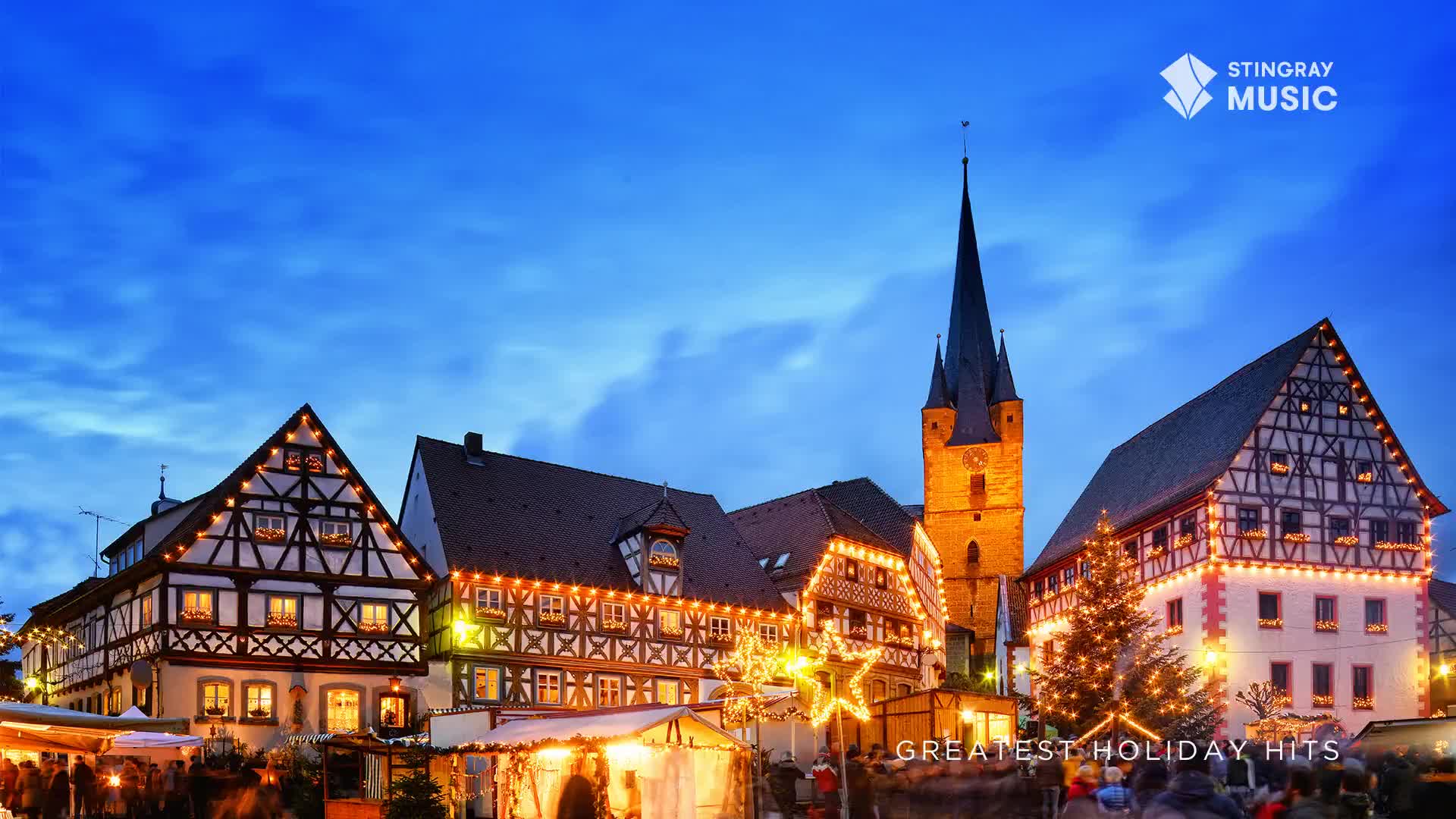 The sky is a deep blue above a town square lit up for the holidays. Buildings with dark roofs and timbered walls are outlined with strings of lights, and a tall church steeple rises in the background.
