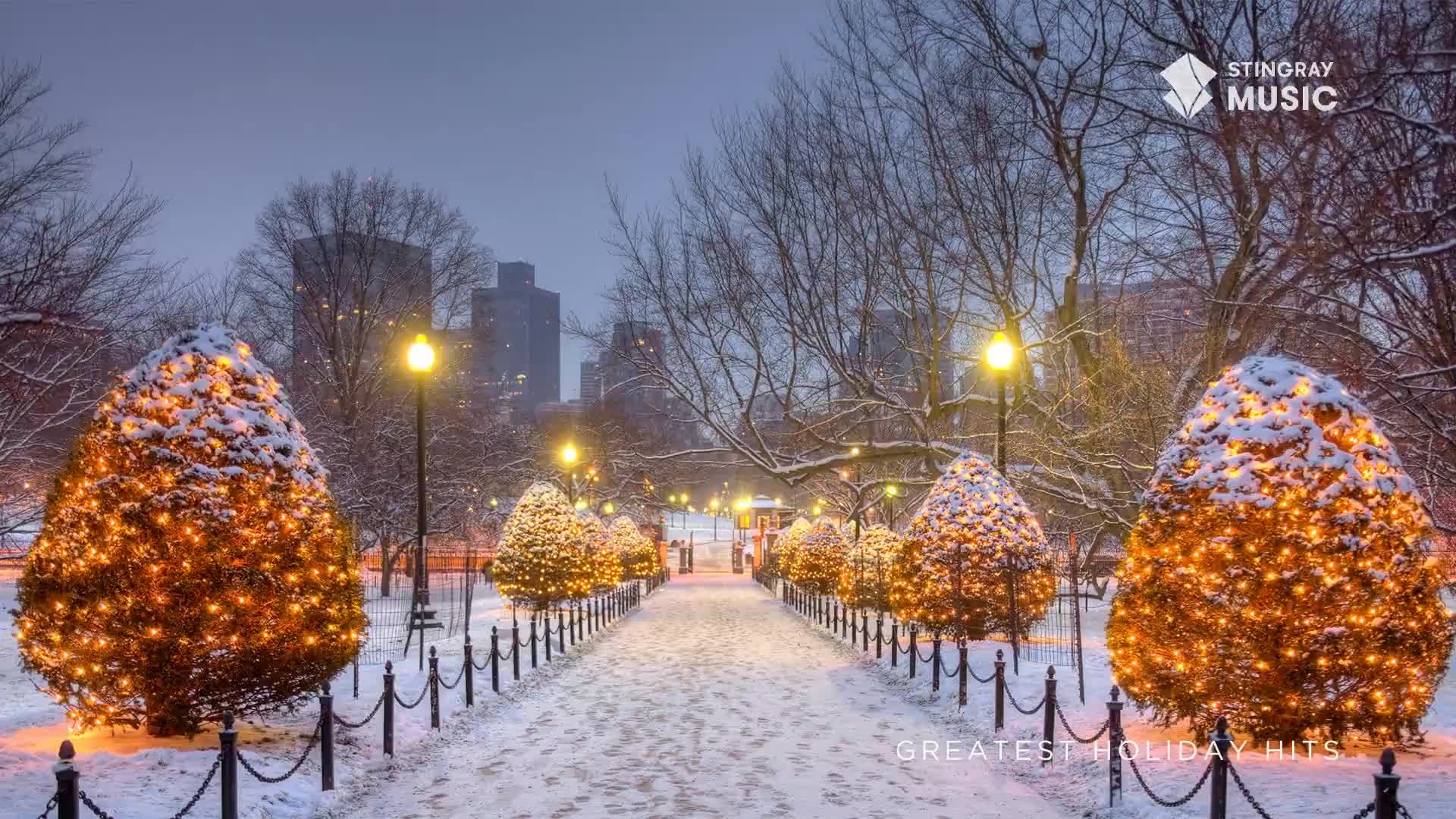 The snow-covered path leads toward the city skyline, lined with glowing, illuminated trees.  It's a scene that feels like a Stingray Holiday Hits commercial from Canada.
