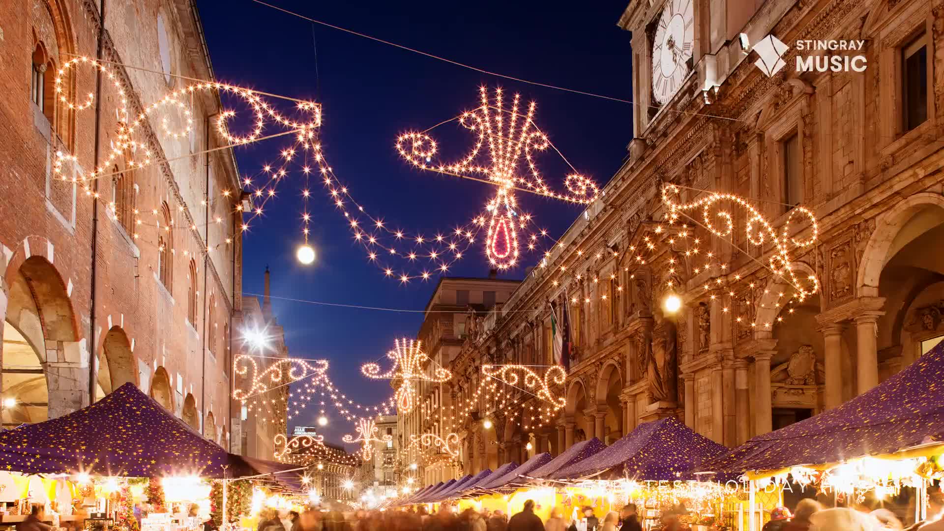 The street is buzzing with people, beneath strings of glowing lights shaped like curlicues and teardrops.  Stalls line the way, their purple canopies adding to the festive atmosphere, likely playing some Stingray Holiday Hits from Canada.

