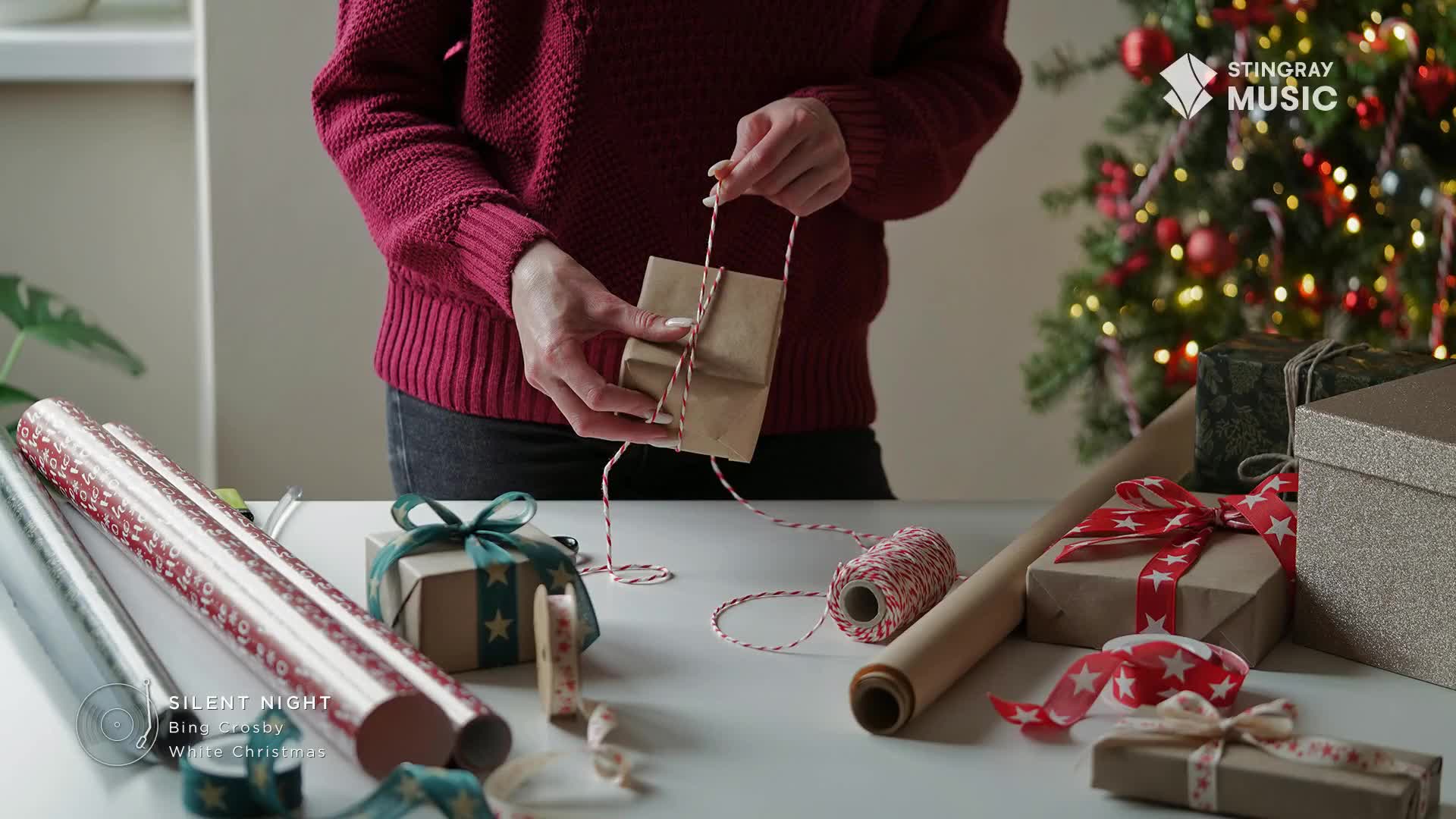 A person in a burgundy sweater is carefully tying a small gift with red and white twine. The Stingray Holiday Hits logo is visible, and the scene is set with a Christmas tree in the background.
