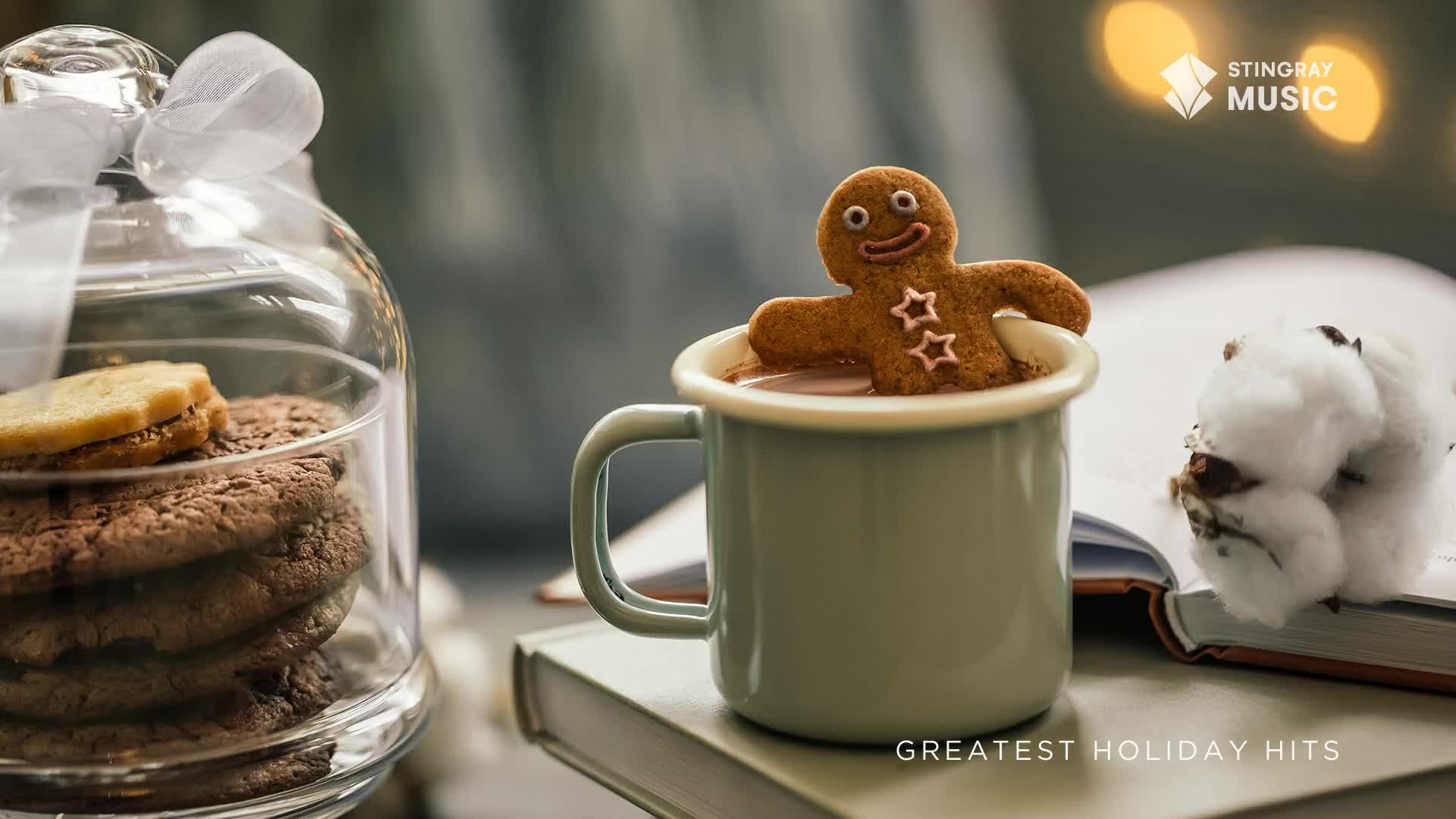 A gingerbread man, with a big smile, is taking a dip in a mug of hot chocolate. Beside it, a stack of cookies sits under a glass dome, ready for a cozy night, perhaps listening to Stingray's Holiday Hits.
