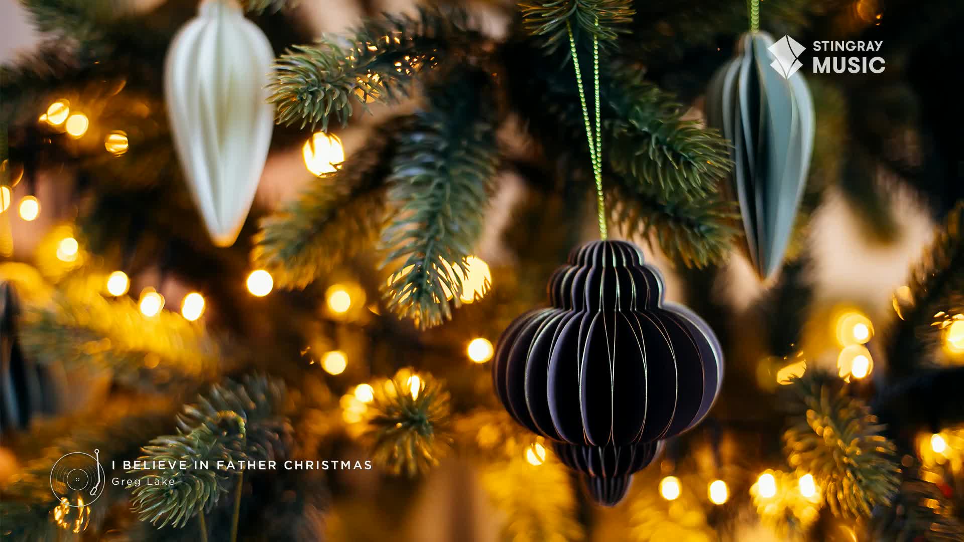 A dark, layered ornament hangs from a Christmas tree, its shape catching the light. Tiny golden lights twinkle in the background, a festive backdrop for Canada's Stingray Holiday Hits.
