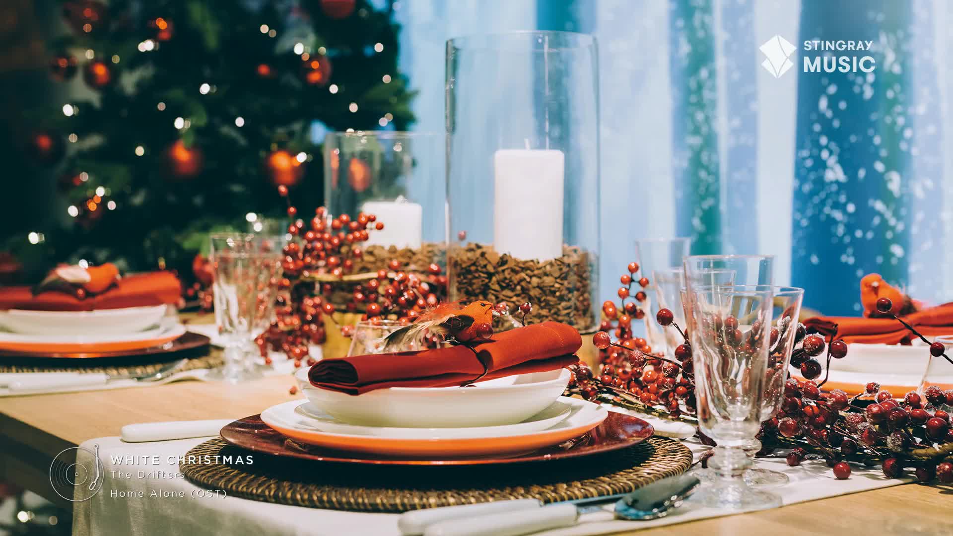 The table is set for a holiday meal, with plates and folded red napkins arranged on woven placemats. Tall glass vases hold candles and berries, and a Christmas tree with twinkling lights is in the background, making it feel like a Stingray Holiday Hits scene from Canada.
