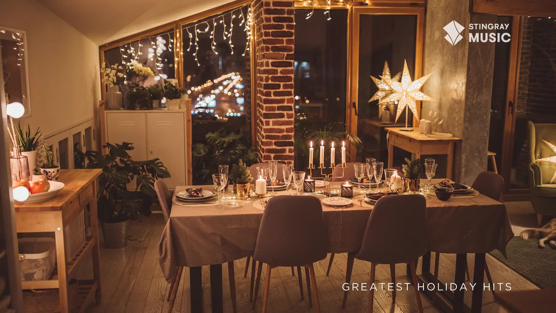 Candlelight flickers across a long table set for a holiday meal, with a backdrop of twinkling lights visible through the windows. The scene feels warm, inviting, and ready for a cozy evening, perhaps with Stingray Holiday Hits playing in the background.

