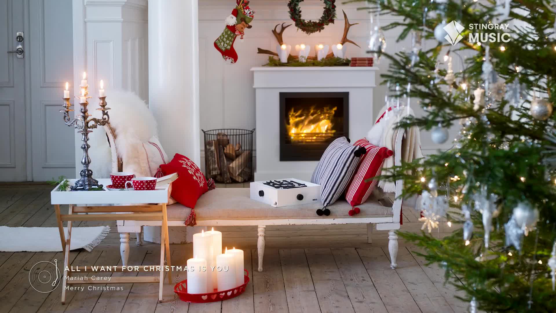 A cozy scene unfolds with a lit fireplace, a decorated Christmas tree, and a record player on a bench. The Stingray Holiday Hits logo is visible, and the song "All I Want for Christmas Is You" by Mariah Carey is playing.
