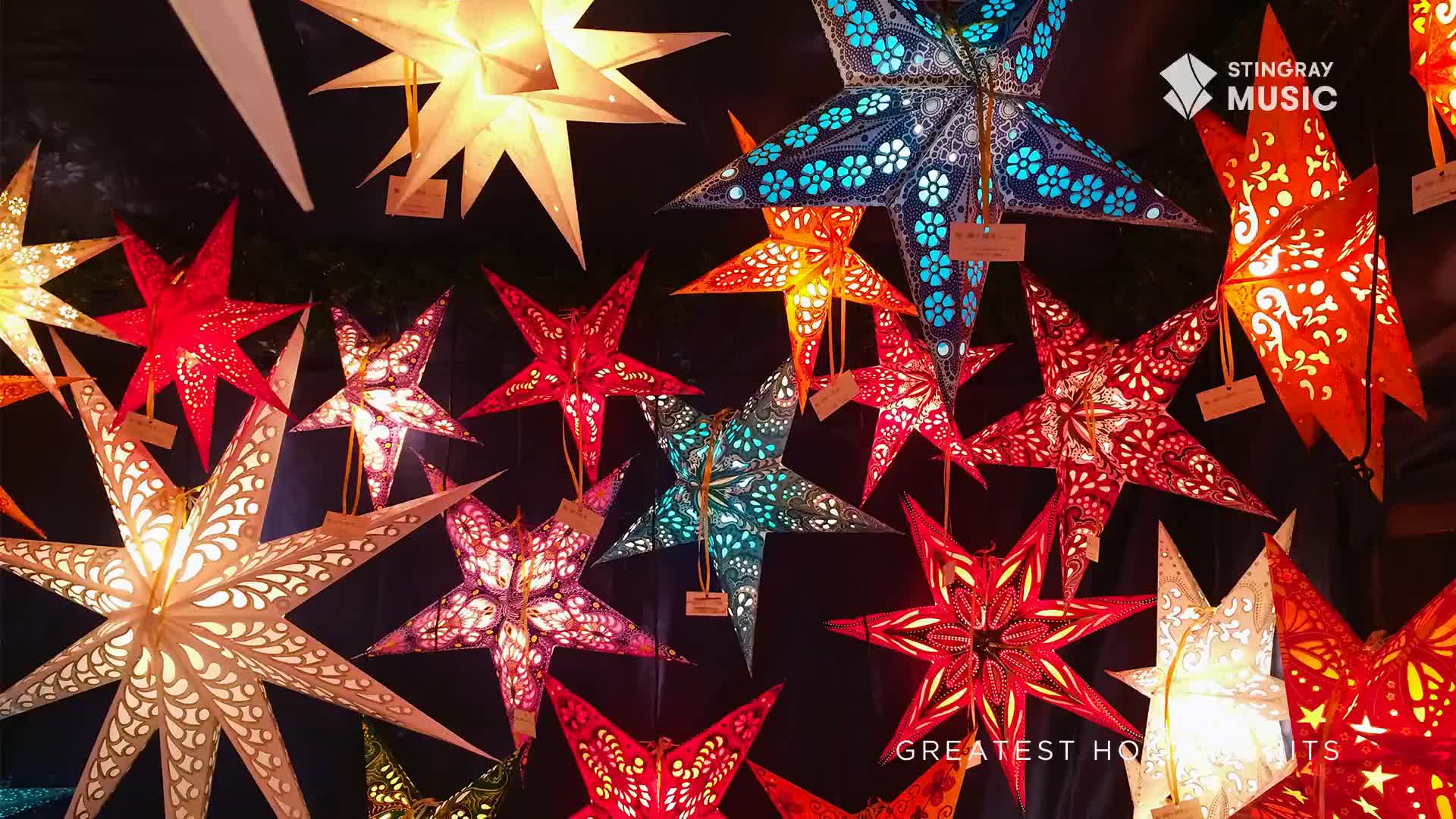 Paper stars in a variety of colors and patterns are suspended, glowing warmly against a dark backdrop. The Stingray Holiday Hits logo is visible, suggesting a festive Canadian atmosphere.
