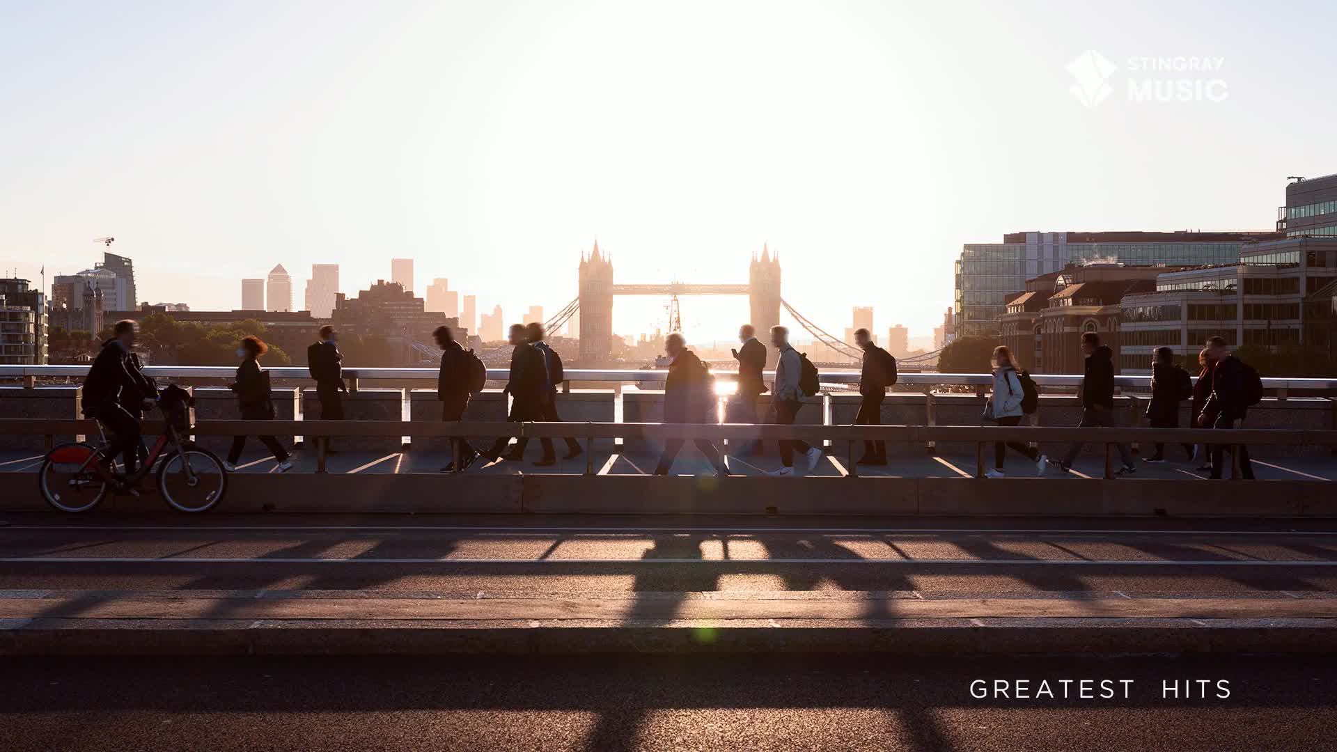 A cyclist on a red bike glides past a line of pedestrians crossing a bridge. The sun casts long shadows across the road as the city skyline, including Tower Bridge, glows in the background.