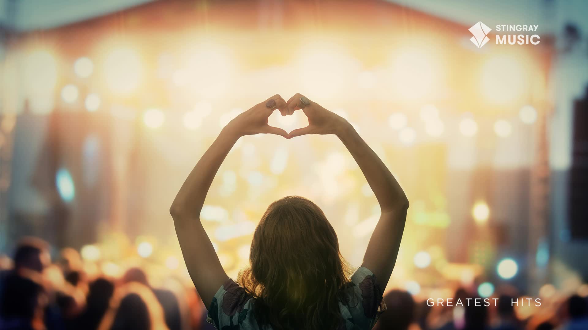 A woman in the crowd forms a heart shape with her hands, her arms raised towards a brightly lit stage. The air buzzes with anticipation for the Stingray Holiday Hits concert.