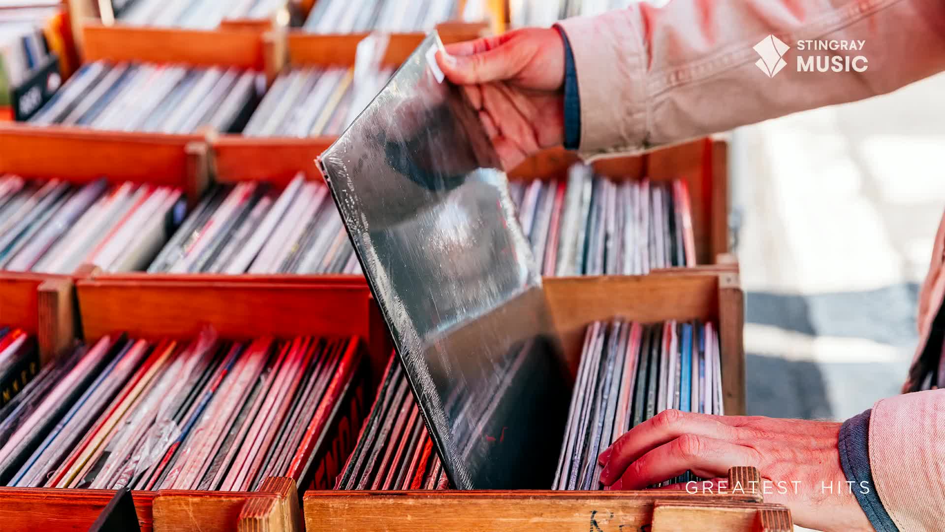 A hand pulls a shiny record from a wooden crate overflowing with albums. Rows of colourful spines, likely classic Canadian hits, fill the crates.
