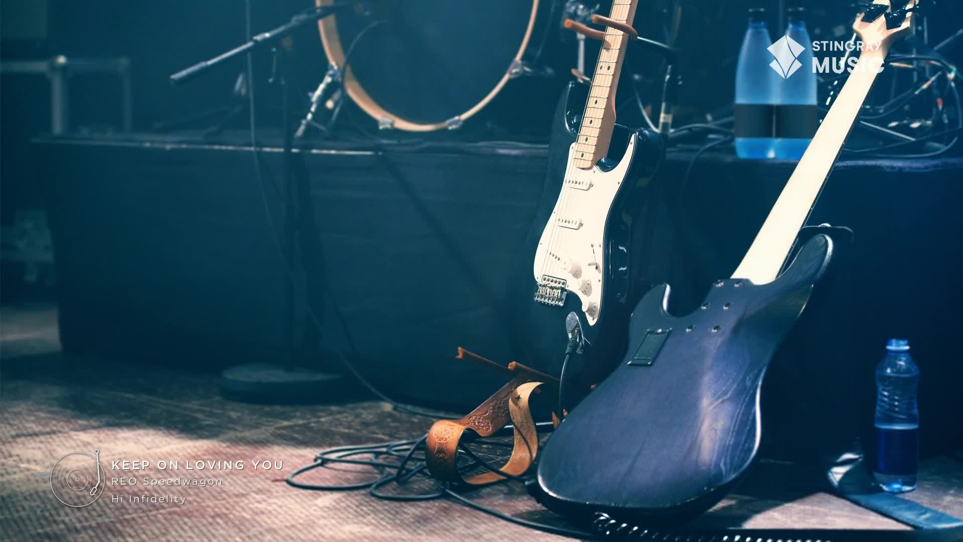 Two electric guitars lean against a dark amp, ready for the next set. A drum kit sits behind them, and bottles of Stingray Holiday Hits water are stacked nearby.