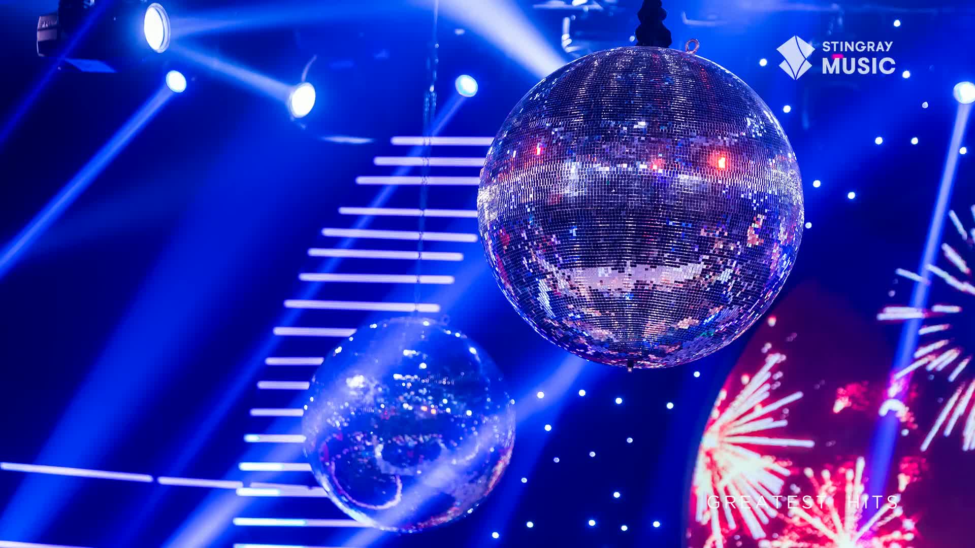 Two large disco balls hang suspended, catching the bright blue stage lights. The larger one, in the foreground, is a shimmering sphere of mirrors, reflecting the vibrant beams.
