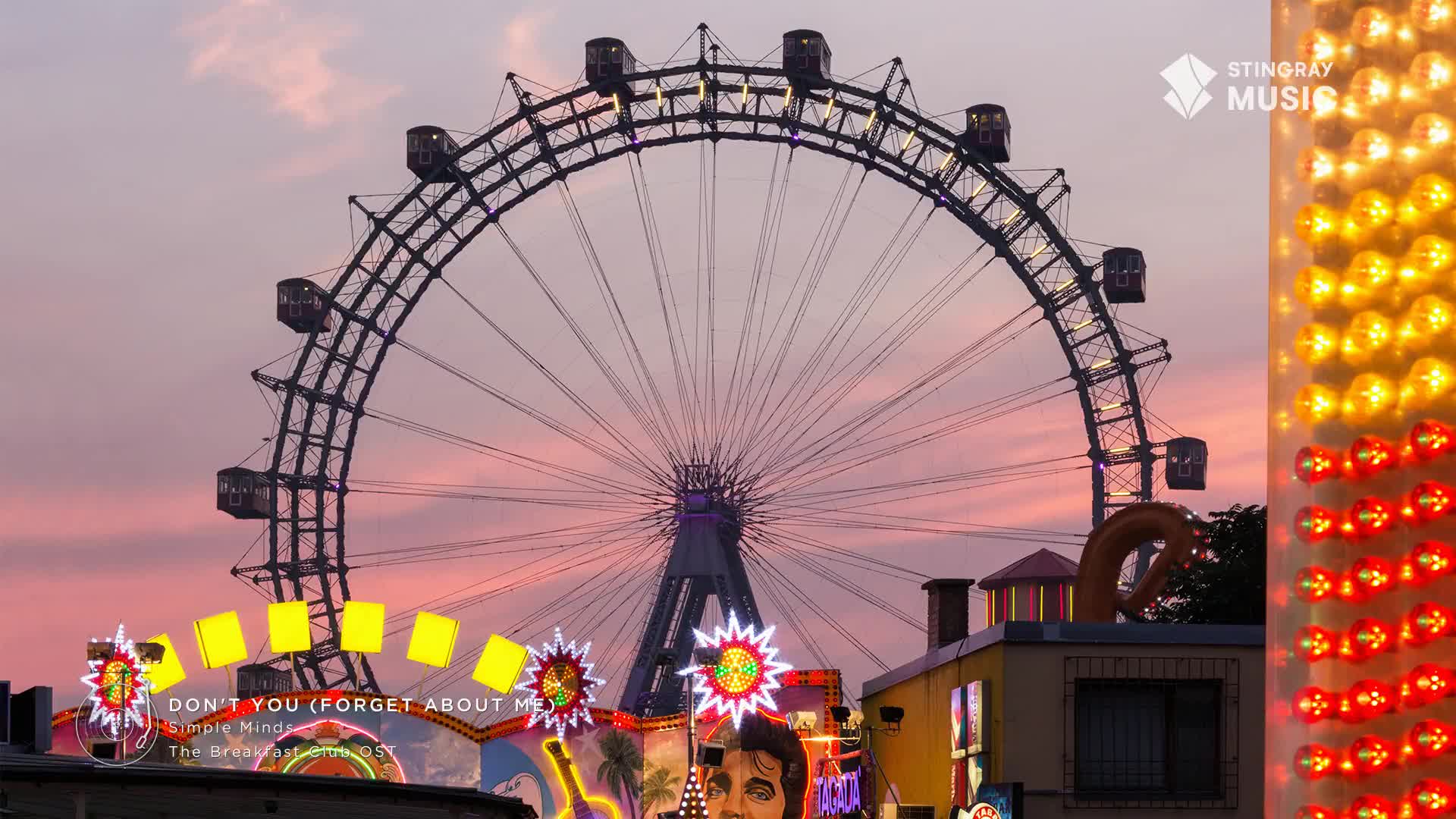 The Ferris wheel's gondolas slowly ascend against a pastel sky, while the bright lights of a nearby attraction pulse with energy. A banner displays "Don't You (Forget About Me)" by Simple Minds, a classic from the Stingray Holiday Hits collection.