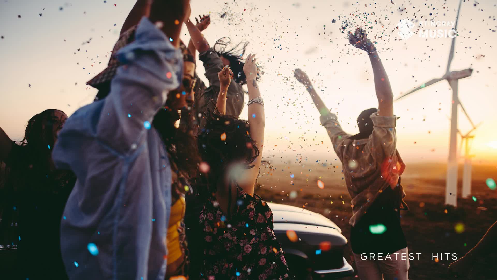 Confetti rains down on a group of friends celebrating near a car, their arms raised in the air. Wind turbines stand tall in the background as the sun sets over the Canadian landscape.