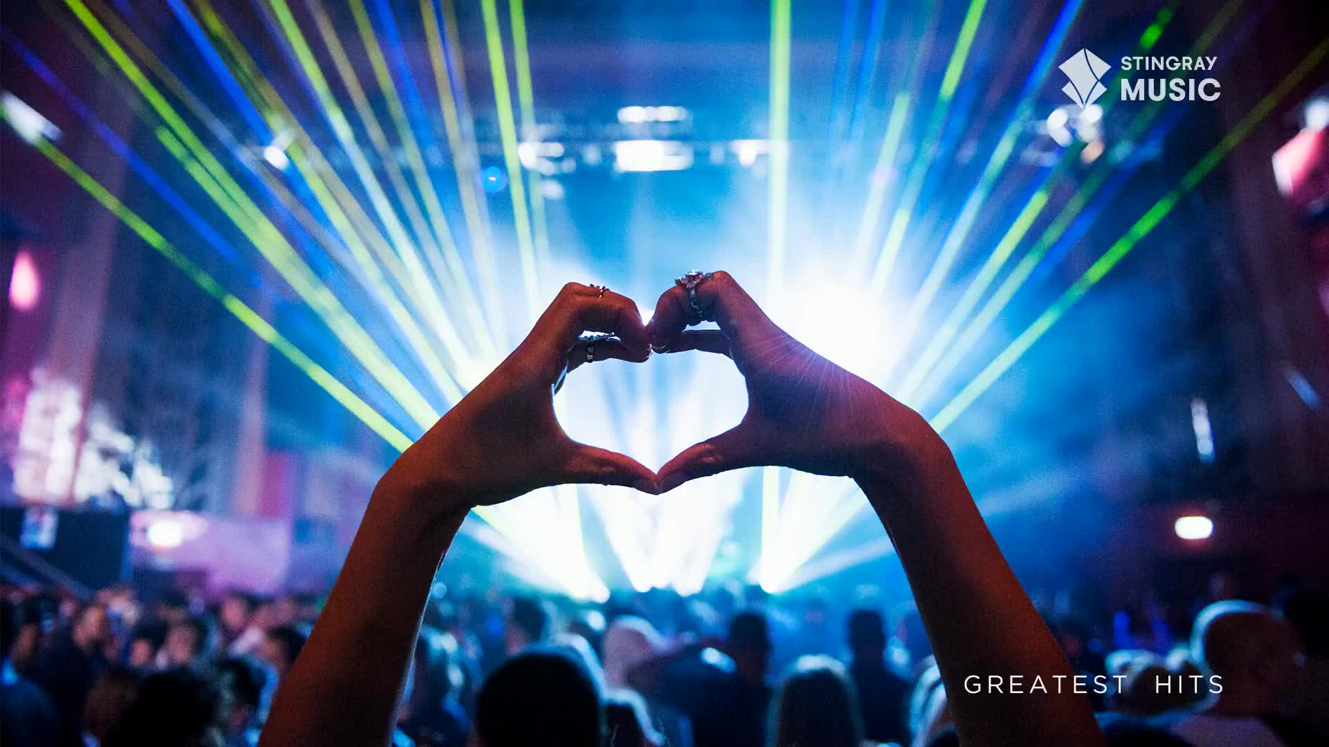 Hands form a heart shape against a backdrop of vibrant blue and green laser lights. A crowd of people is visible below, enjoying the show.