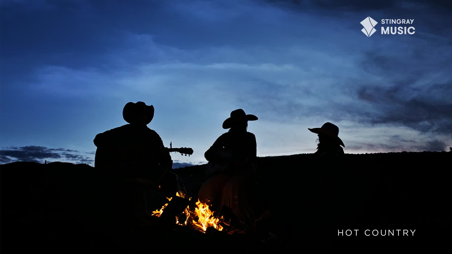 Under a darkening sky, three figures in cowboy hats sit around a crackling fire. The silhouettes of guitars are visible, promising a night of music under the Canadian stars for Stingray Hot Country.
