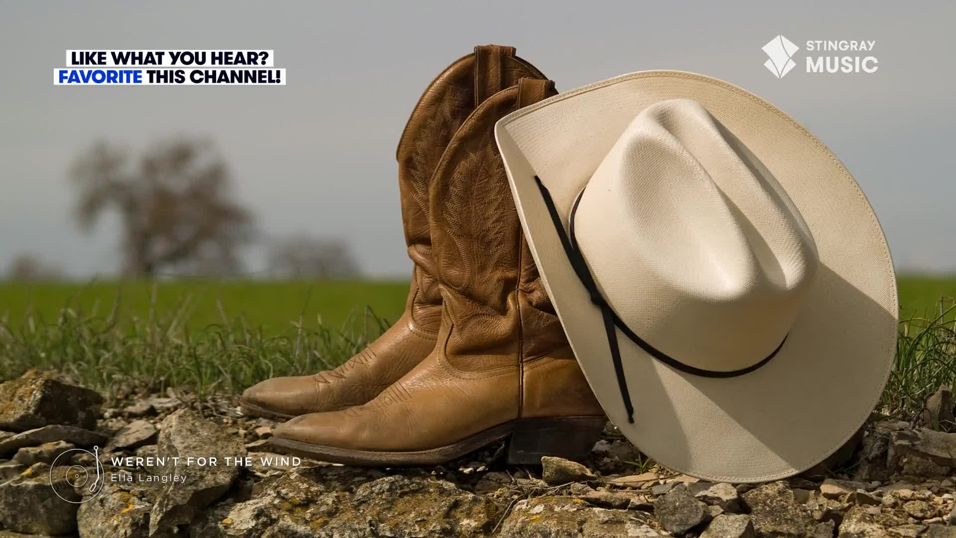 A pair of well-worn cowboy boots and a white hat rest on a stone wall. The Stingray Hot Country logo is visible, and the song "Weren't For The Wind" is playing.
