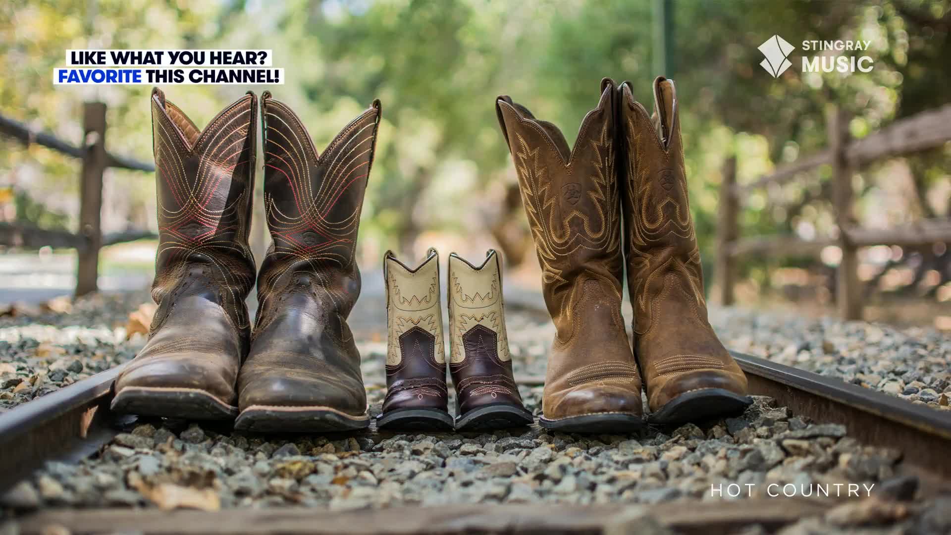 Four pairs of cowboy boots stand on the tracks, two adult pairs flanking two smaller ones. The scene, backed by a wooden fence and soft green trees, is reminiscent of a Stingray Hot Country promotion.
