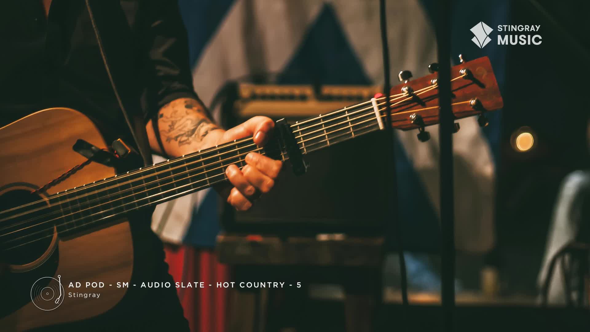 A musician strums an acoustic guitar, the capo clamped on the frets. The camera focuses on the instrument and the hand with a tattoo, likely a performance for Stingray Hot Country.
