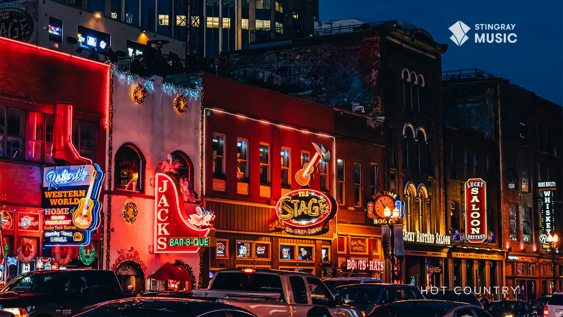 Neon signs blaze, advertising bars and restaurants along a street at night. Cars crawl forward, their headlights reflecting off the wet pavement.

