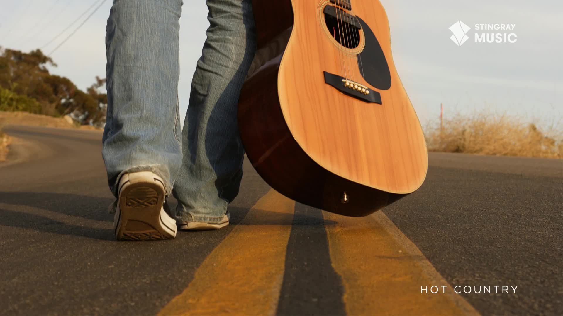 A person in jeans and white sneakers walks along a road, a guitar cradled in their arm. The Stingray Hot Country logo is visible, and the scene feels like a drive through Canada.
