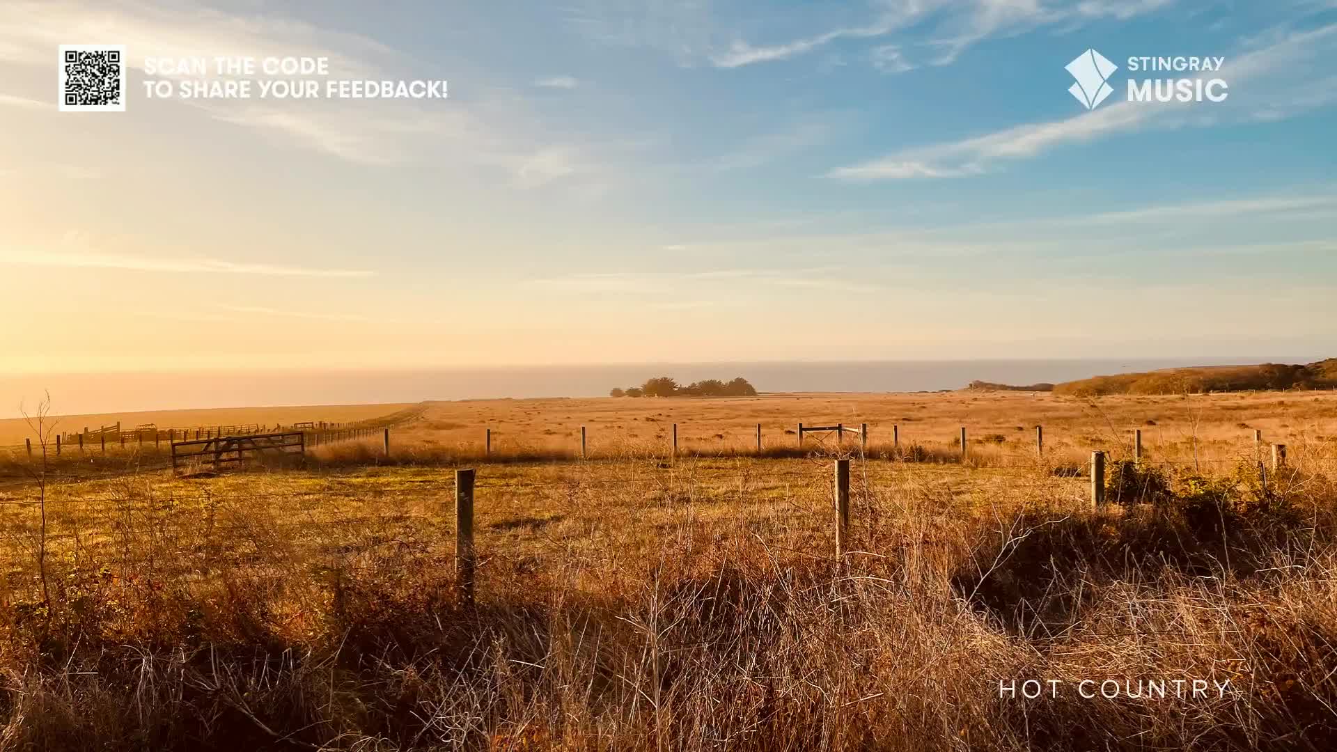 The sun is setting over the ocean, casting a warm glow across the fields. A weathered fence runs along the edge of the dry, golden grass.
