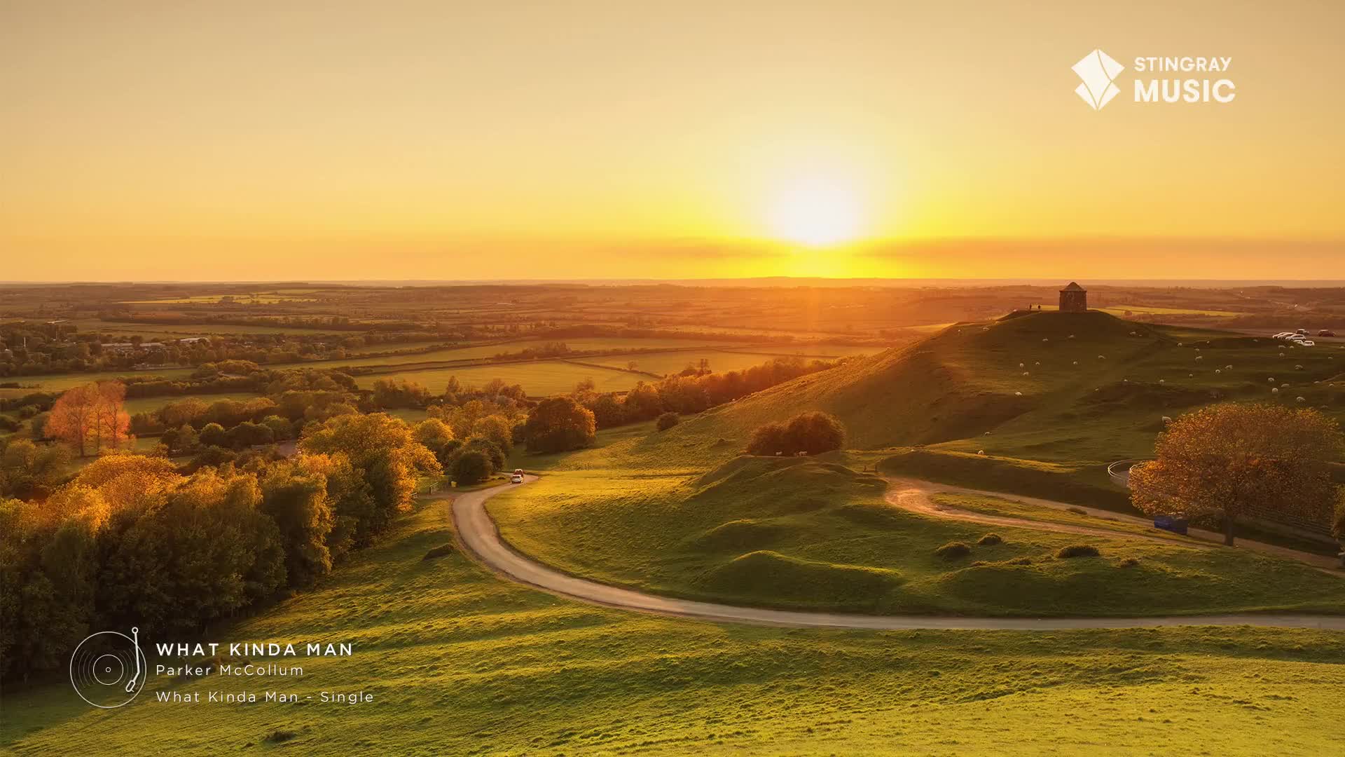 A winding road curves through the green hills, leading toward a building silhouetted against the setting sun. The sky is ablaze with orange and yellow, casting a warm glow over the landscape.
