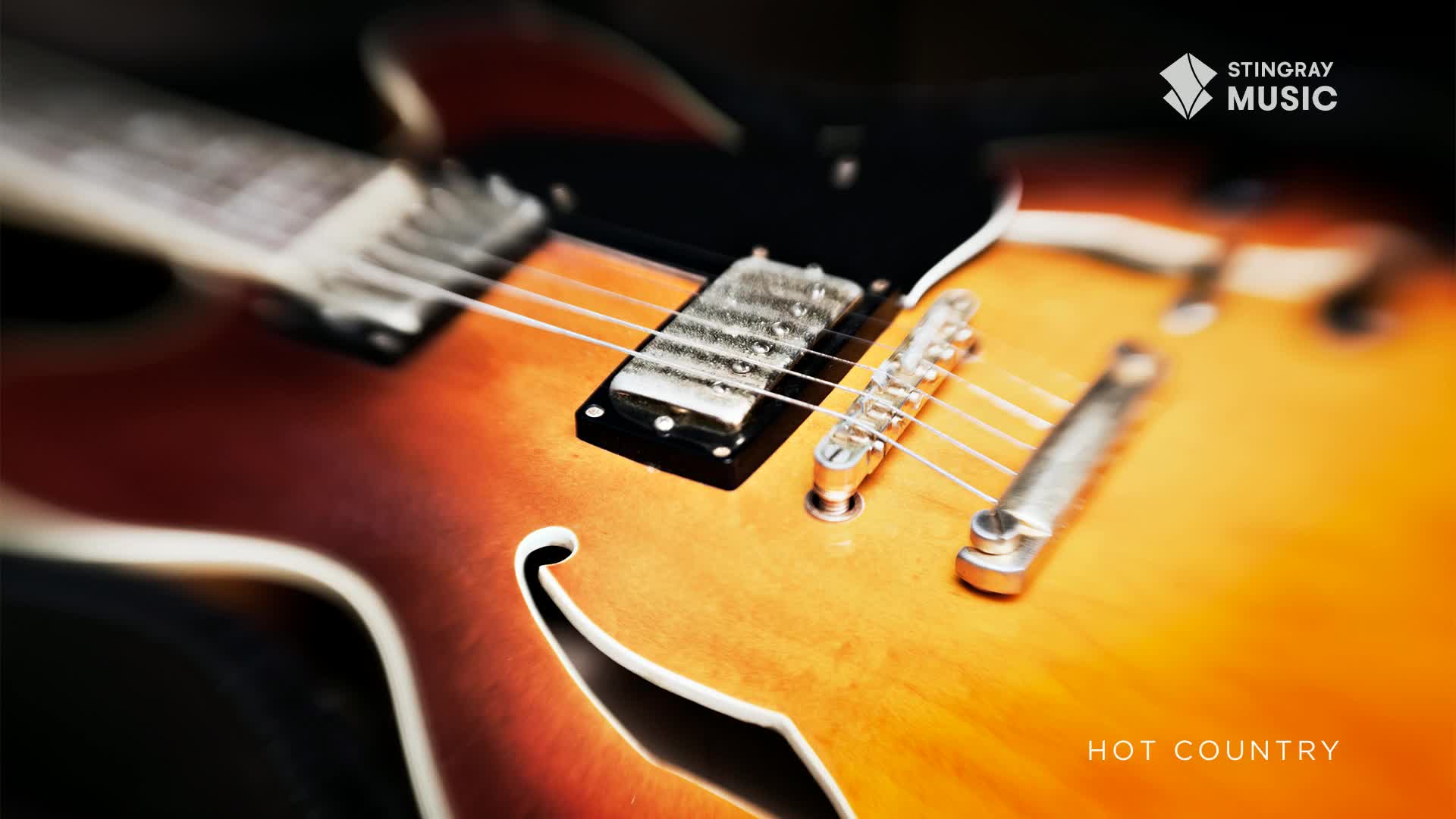 The body of a sunburst electric guitar is in sharp focus, its strings gleaming.  "Hot Country" is written below the instrument, and the Stingray Music logo is in the corner.
