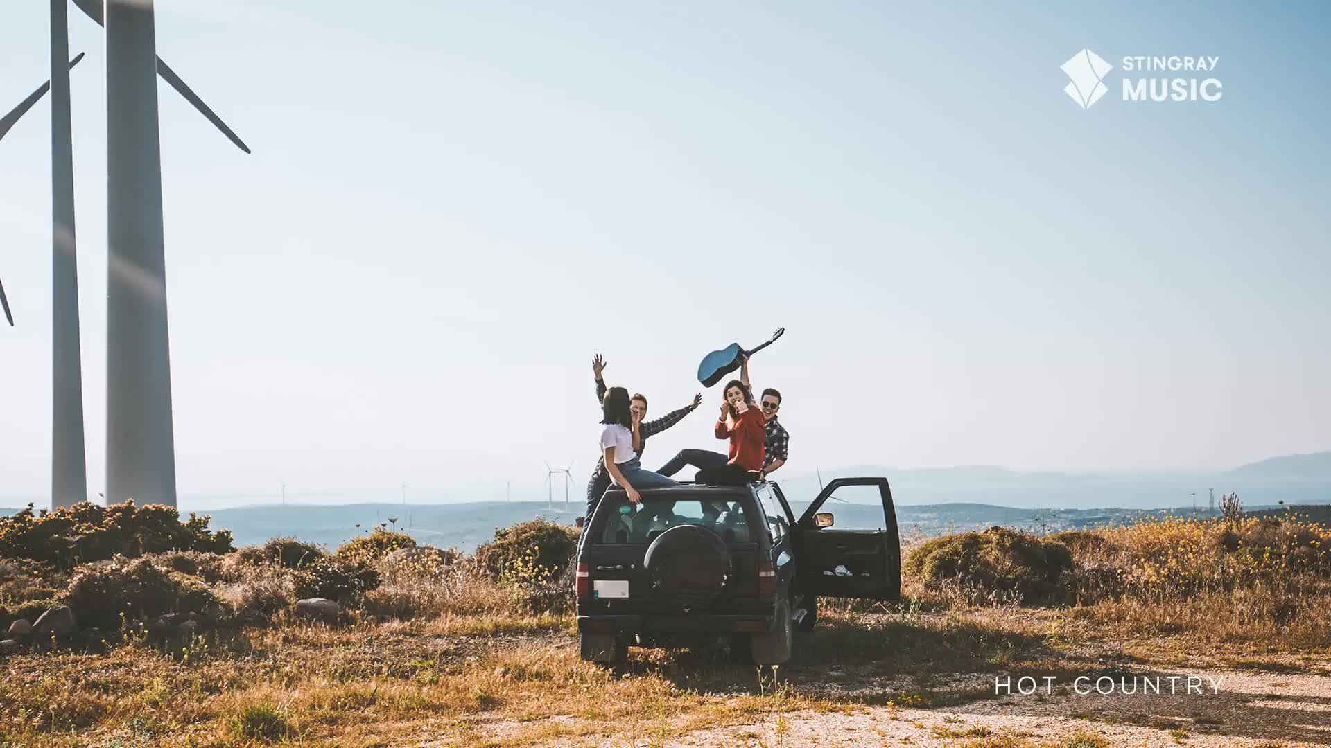 A group of friends sits on top of a dark-colored SUV, one holding a guitar high in the air. Wind turbines tower in the background, and the scene feels like a perfect day for some Hot Country music.
