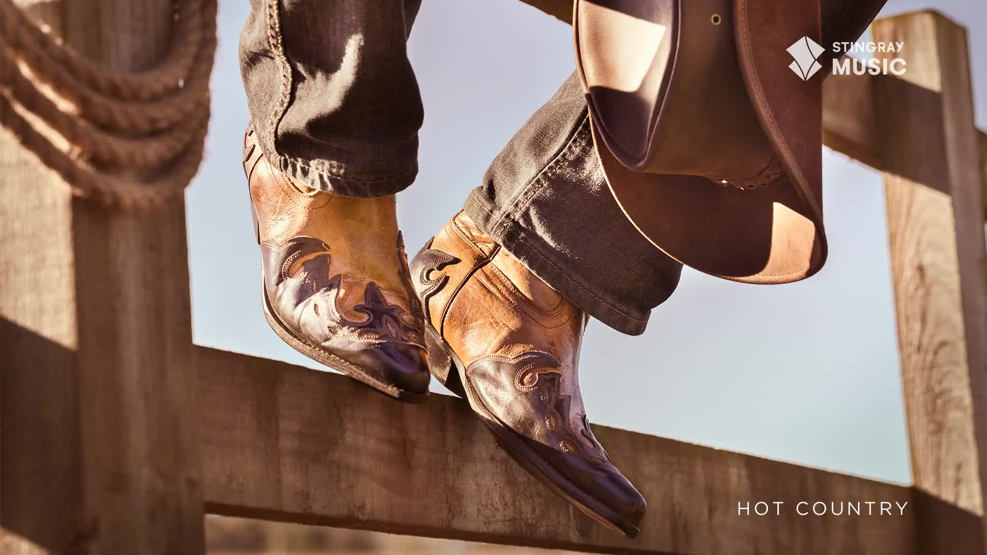 A pair of cowboy boots, brown and tan with intricate designs, rest on a wooden fence. The boots, and the Stingray Hot Country branding, evoke a feeling of the Canadian countryside.
