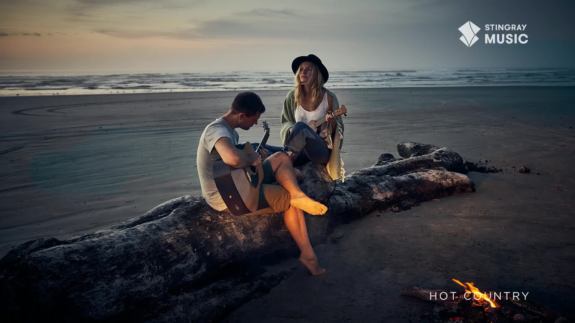 On a beach, a man and a woman sit on a log, strumming acoustic guitars. The Stingray Hot Country logo is visible, and a small fire crackles nearby.
