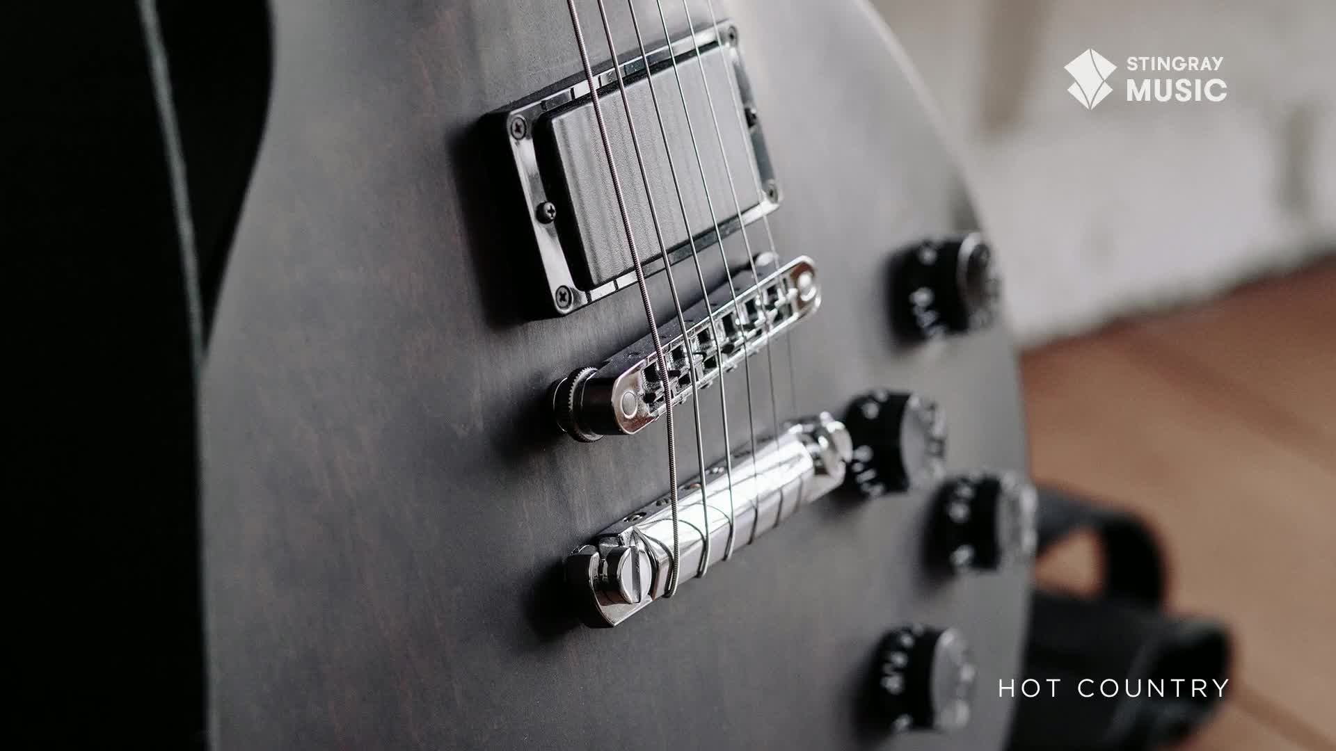 A matte black electric guitar, likely for Stingray Hot Country in Canada, is in sharp focus. The strings gleam against the dark body, and the chrome bridge catches the light.
