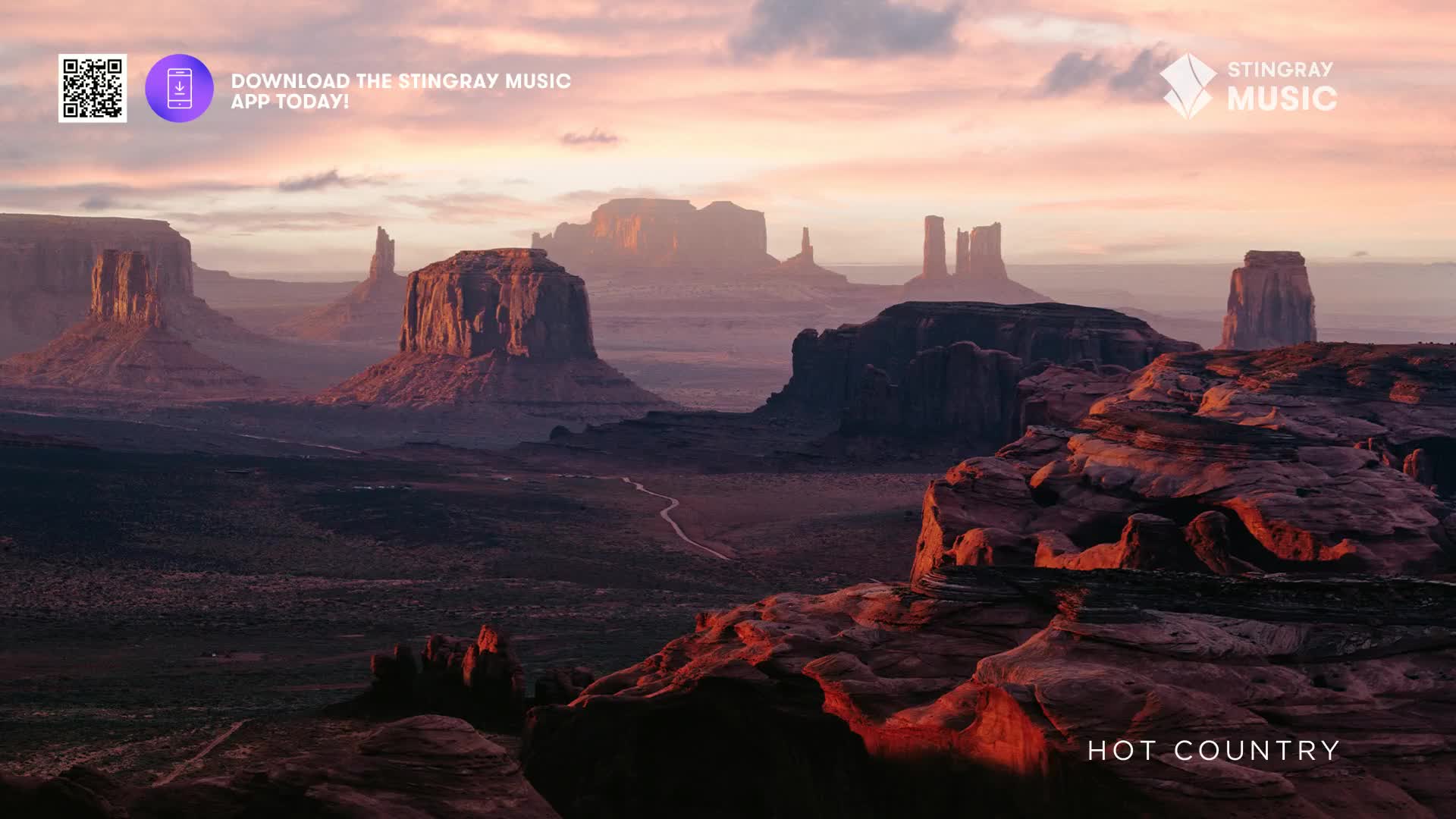 The sun casts a warm glow on the towering rock formations, their shadows stretching across the valley floor. The Stingray Hot Country logo sits at the bottom, promising the soundtrack to this vast landscape.
