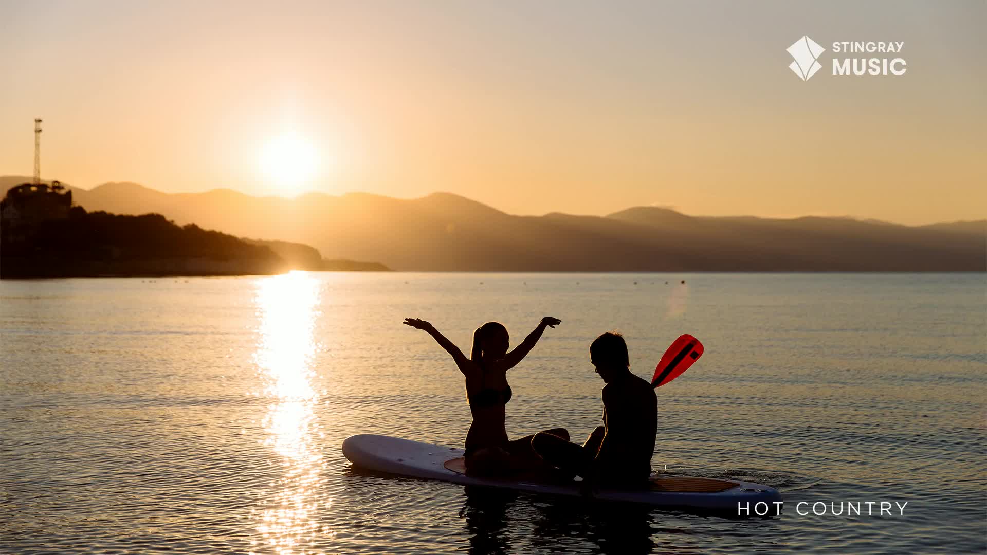 The sun sets over the water as a couple sits on a paddleboard. One person has their arms raised in the air, silhouetted against the glowing sky, while the other holds a paddle.

