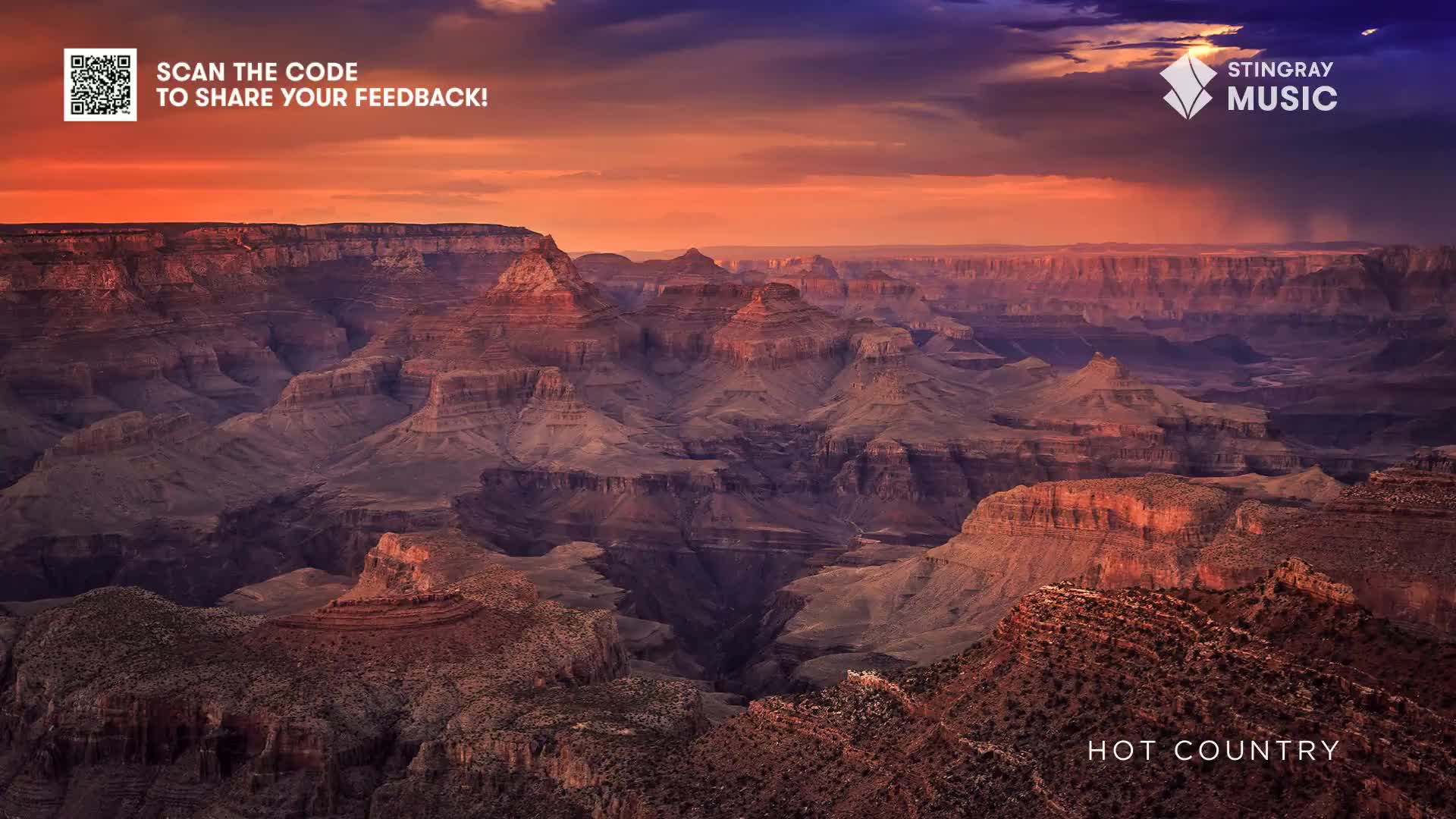 The vast canyon stretches out, its layered rock formations catching the warm hues of the sunset. Above, the sky is a dramatic blend of orange and purple, setting the stage for Stingray Hot Country.
