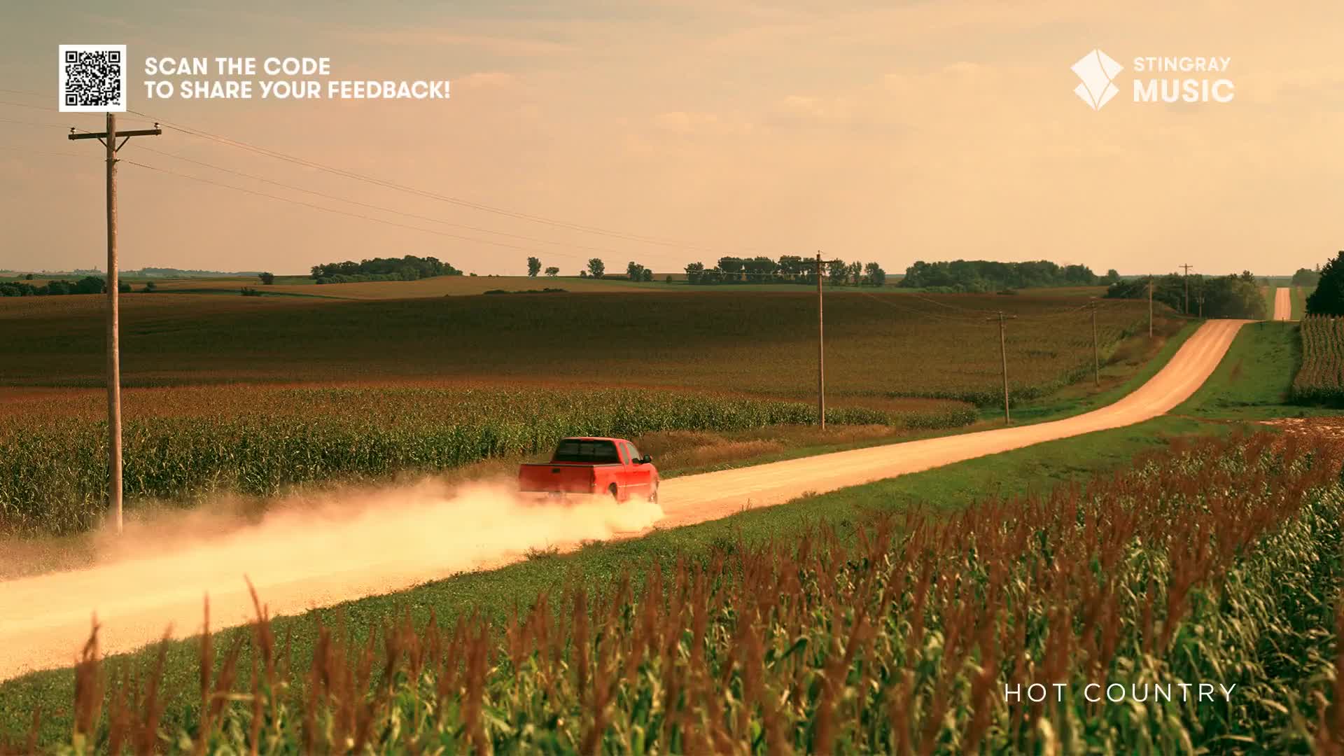 A red pickup truck kicks up a cloud of dust as it speeds down a long, gravel road. Fields of corn flank the road, and the sky is a warm, hazy color.
