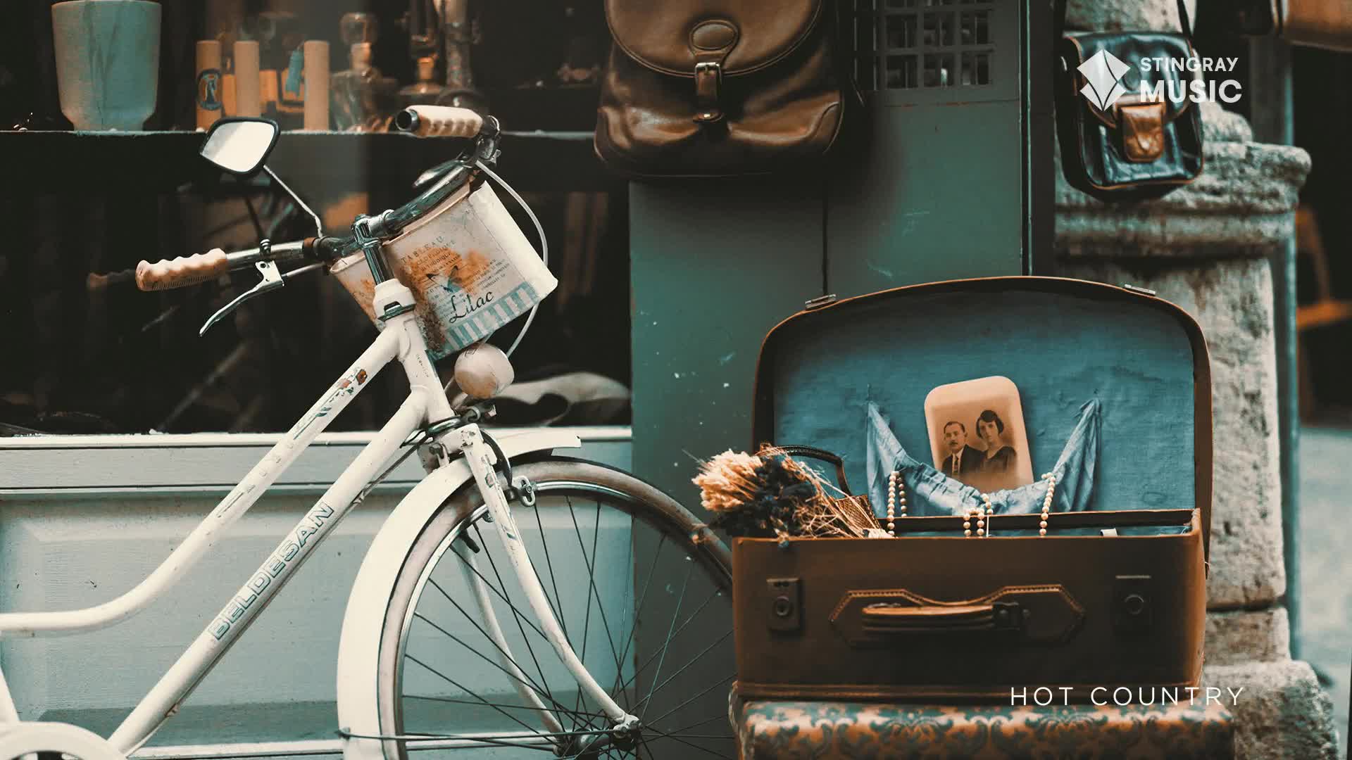 A vintage white bicycle leans against a wall, its basket filled with a floral-patterned fabric. Beside it, an open suitcase displays a framed photograph and a string of pearls, evoking the feel of Stingray Hot Country in Canada.
