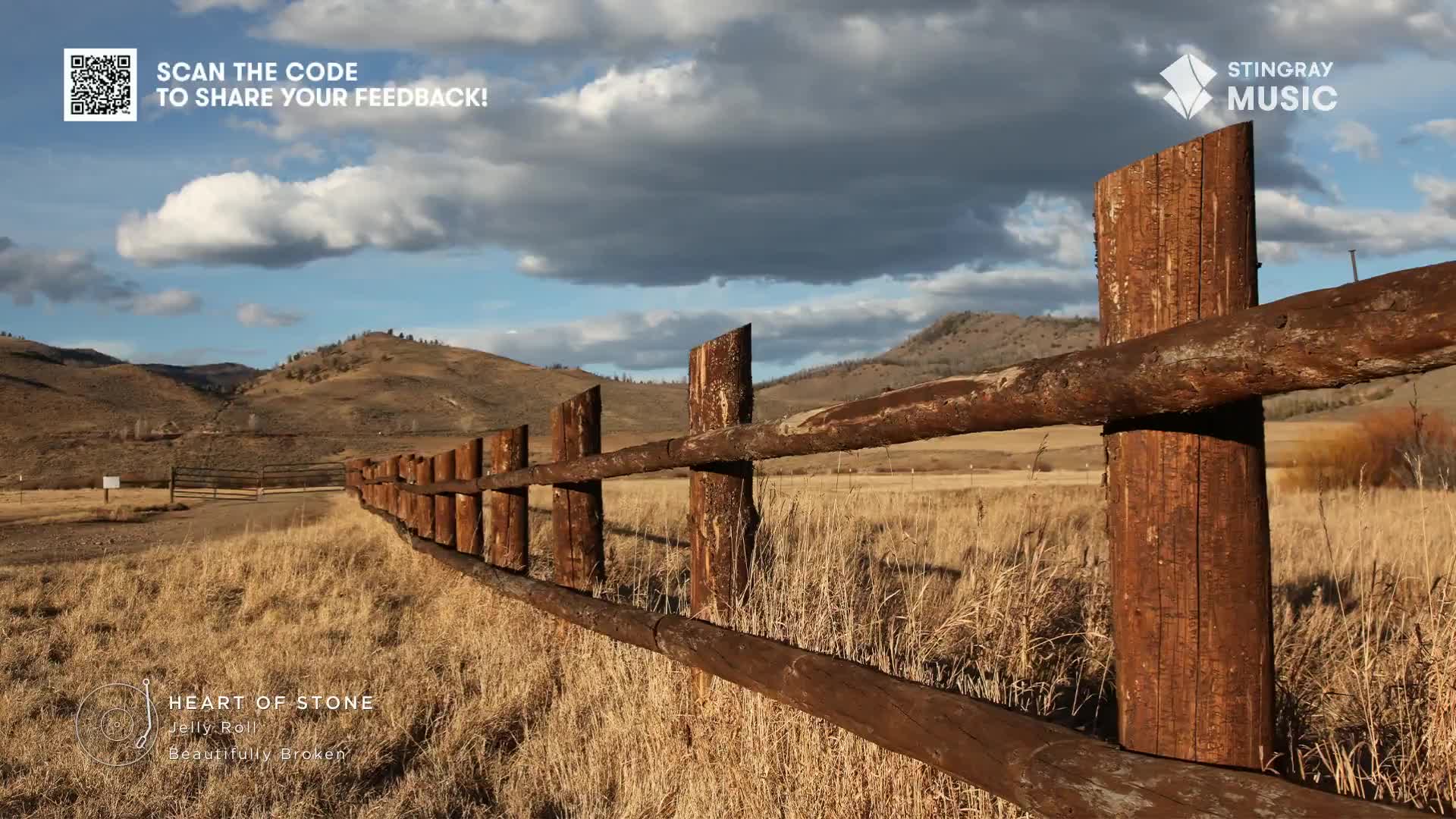 A weathered wooden fence stretches across a field of dry grass, leading the eye towards the distant hills. The sky is a mix of blue and cloud, and the air feels crisp, like a Stingray Hot Country song on a drive through Canada.
