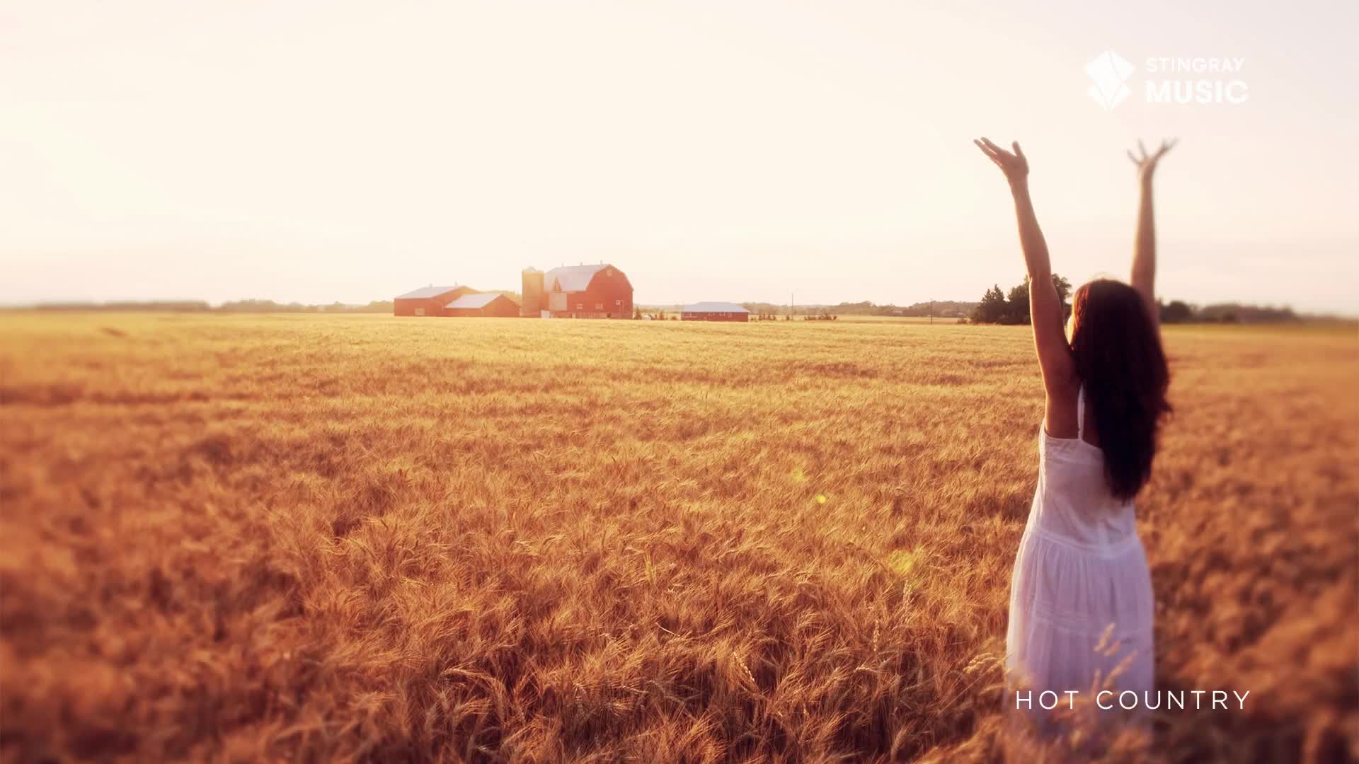 A woman in a white dress stands in a field, arms raised toward the sun. In the distance, a red barn and other farm buildings sit bathed in warm light, a scene that feels like Canada's Stingray Hot Country.
A woman in a white dress stands in a field, arms raised toward the sun. In the distance, a red barn and other farm buildings sit bathed in warm light, a scene that feels like Canada's Stingray Hot Country.