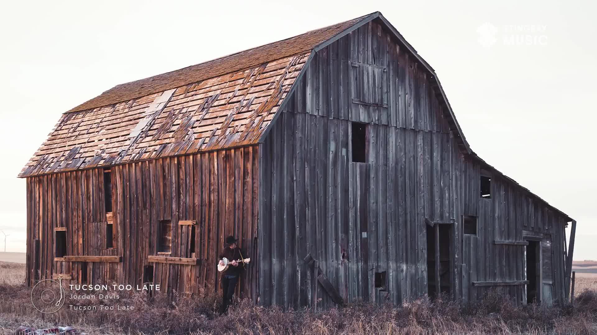 A musician stands in front of a weathered barn, strumming a guitar. The building's wood is faded and worn, with a roof that's seen better days.
A musician stands in front of a weathered barn, strumming a guitar. The building's wood is faded and worn, with a roof that's seen better days.