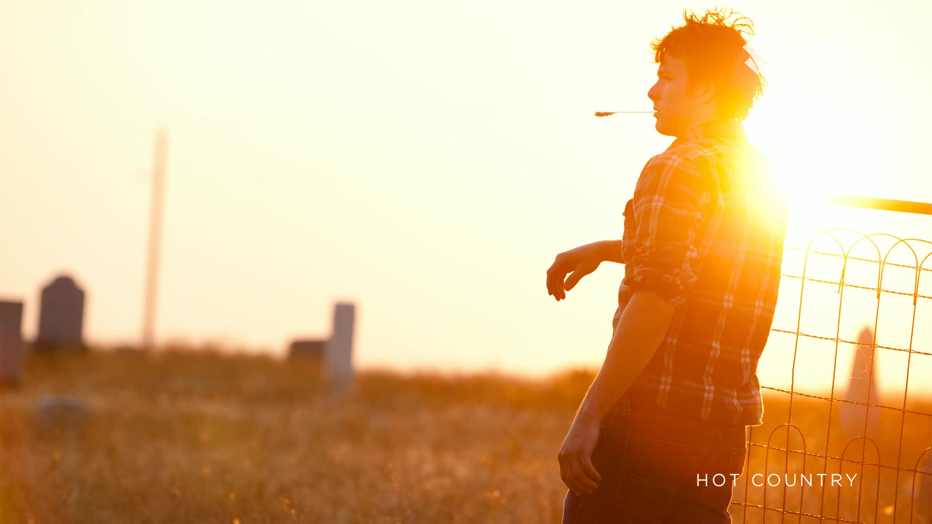 The sun is low and bright, backlighting a young man with a cigarette in his mouth, leaning against a wire fence. Beyond him, a graveyard stands in silhouette against the golden sky, a scene that feels like a still from a Canadian drama.
The sun is low and bright, backlighting a young man with a cigarette in his mouth, leaning against a wire fence. Beyond him, a graveyard stands in silhouette against the golden sky, a scene that feels like a still from a Canadian drama.