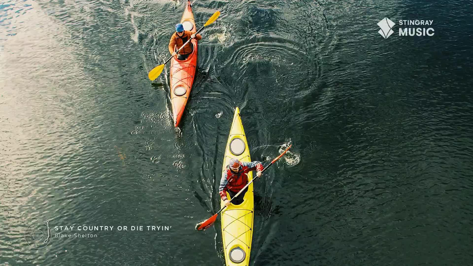 Two kayakers, one in a yellow boat and the other in red, paddle across the dark water, leaving swirling trails behind them. The Stingray Hot Country logo sits in the top right corner, and the song title "Stay Country or Die Tryin'" is printed at the bottom.
Two kayakers, one in a yellow boat and the other in red, paddle across the dark water, leaving swirling trails behind them. The Stingray Hot Country logo sits in the top right corner, and the song title "Stay Country or Die Tryin'" is printed at the bottom.
