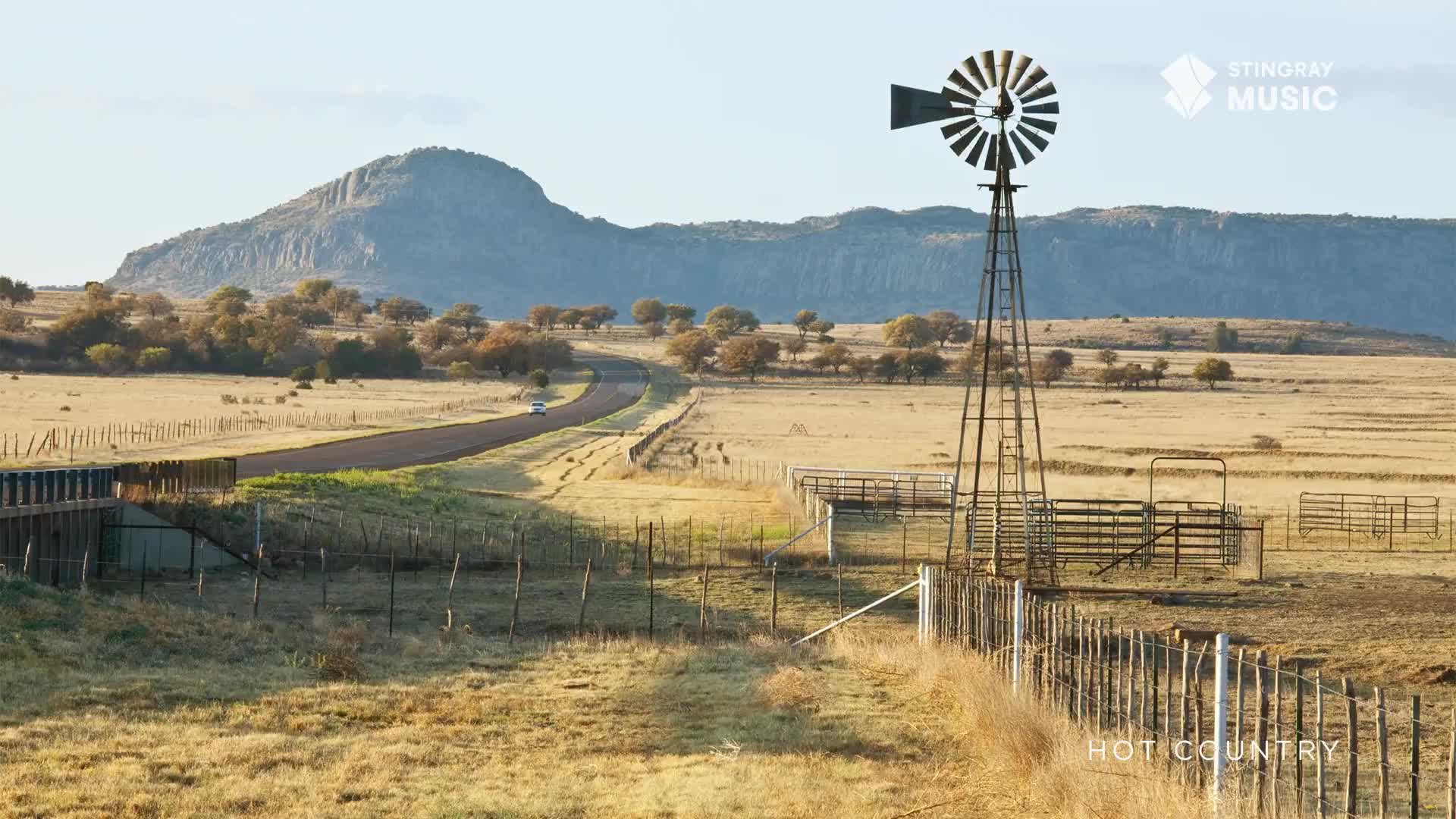 A lone car travels along a winding road, disappearing into the distance. The blades of a windmill stand still against the backdrop of a large, rocky hill.
A lone car travels along a winding road, disappearing into the distance. The blades of a windmill stand still against the backdrop of a large, rocky hill.