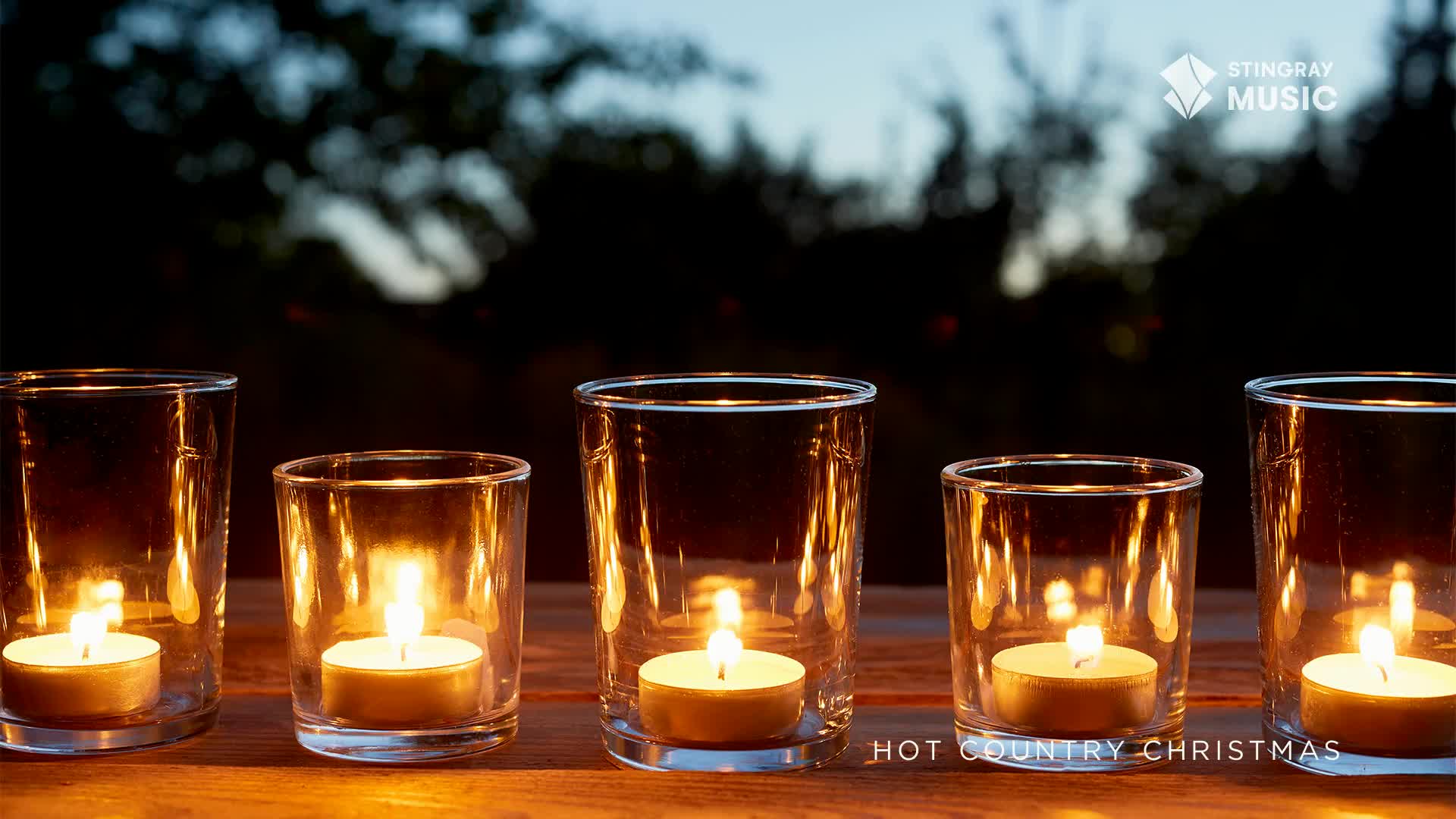 Five small candles flicker inside clear glass holders, casting a warm glow on the wooden surface. The scene is set outdoors, under a dark sky, and the Stingray Hot Country Christmas logo is visible.
Five small candles flicker inside clear glass holders, casting a warm glow on the wooden surface. The scene is set outdoors, under a dark sky, and the Stingray Hot Country Christmas logo is visible.