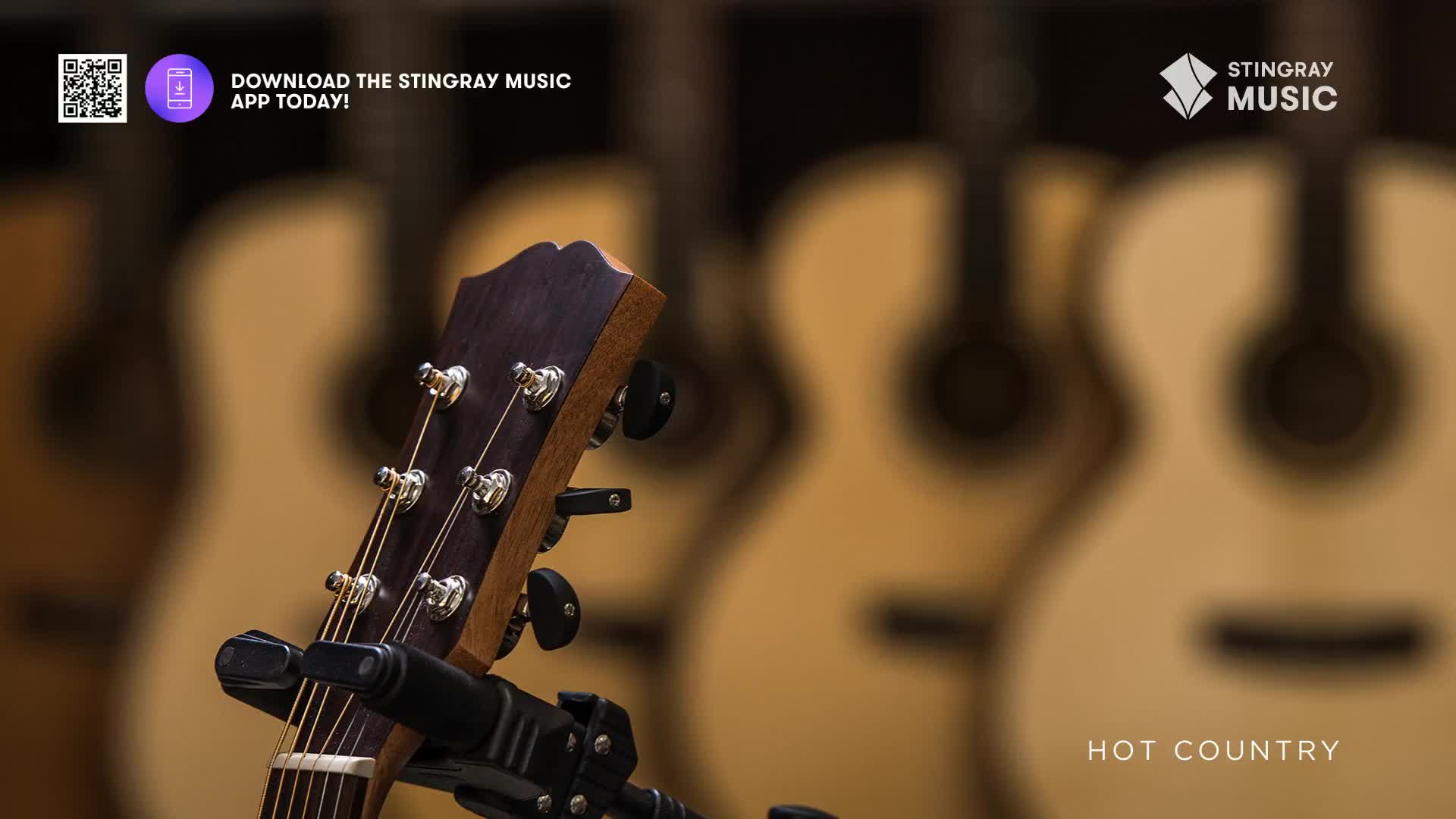 A dark wood guitar headstock with tuning pegs stands front and center, supported by a stand. Behind it, several acoustic guitars with light wood bodies are softly out of focus.