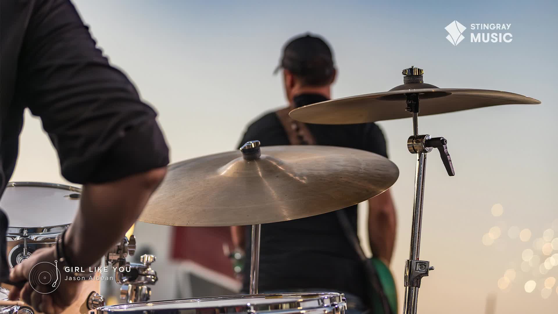 The drummer's arm is raised, ready to strike the cymbals. A band member in a black t-shirt and baseball cap stands further back, facing away.