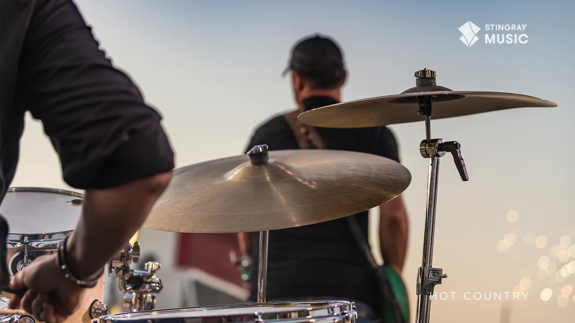 A drummer in a black shirt plays a beat on the cymbals, his sticks a blur against the bright sky. Behind him, a bandmate in a baseball cap waits his turn, the sounds of Stingray Hot Country filling the air.