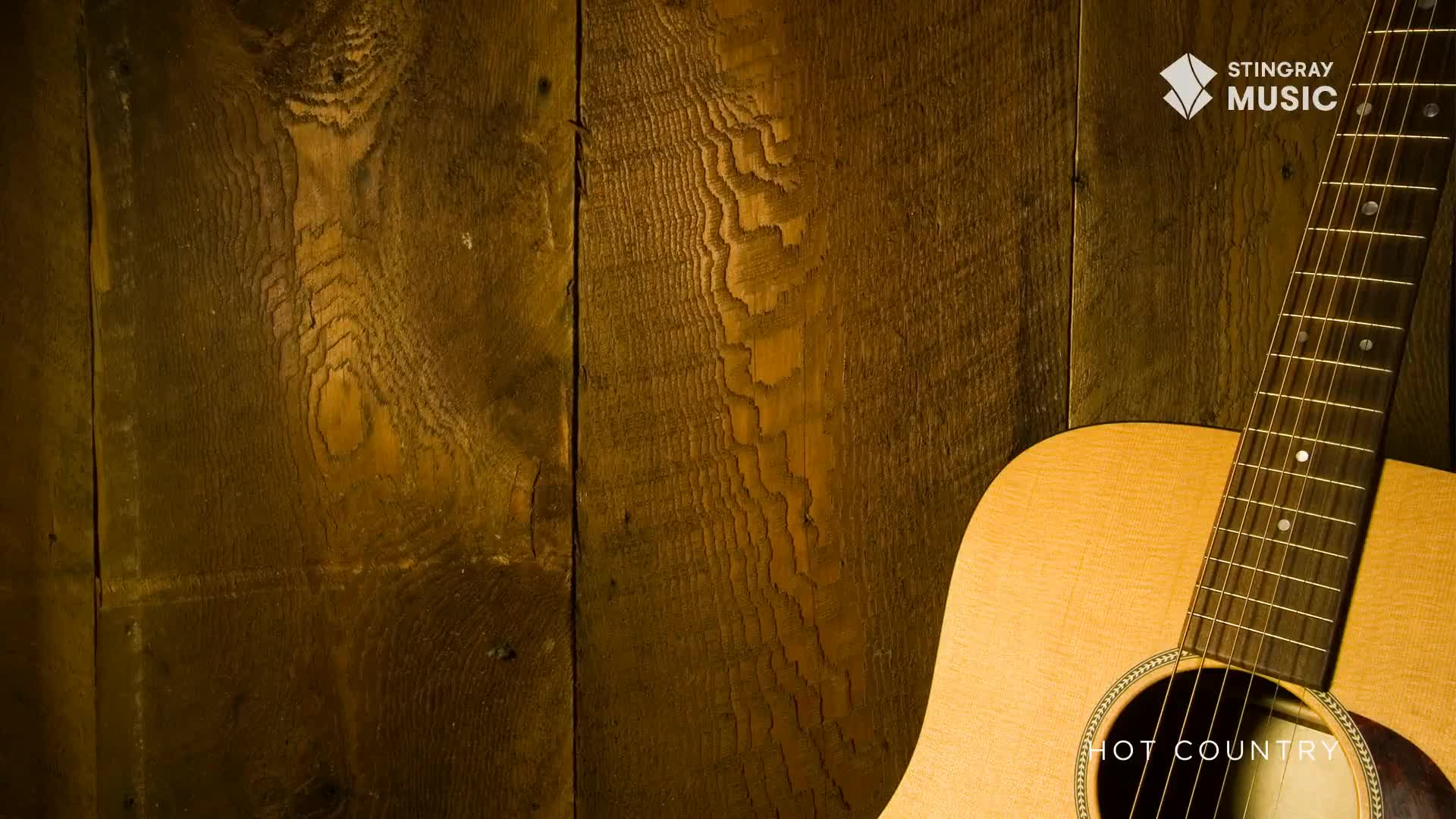 The warm wood grain of a rustic Canadian barn wall serves as a backdrop. A light-colored acoustic guitar rests against it, its fretboard and soundhole clearly visible.